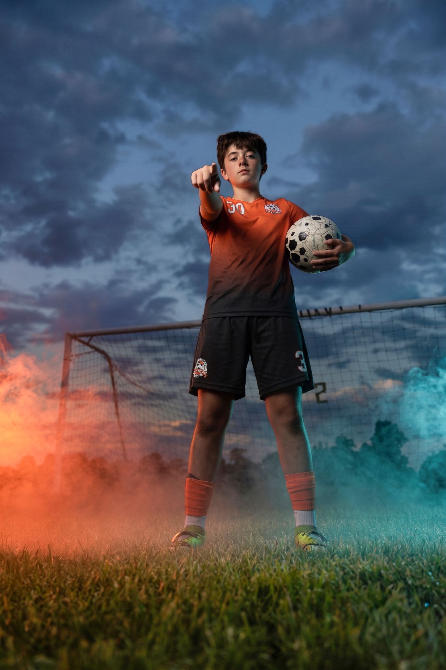 Young soccer player in orange uniform holds ball, pointing forward, surrounded by colorful smoke on field at dusk.