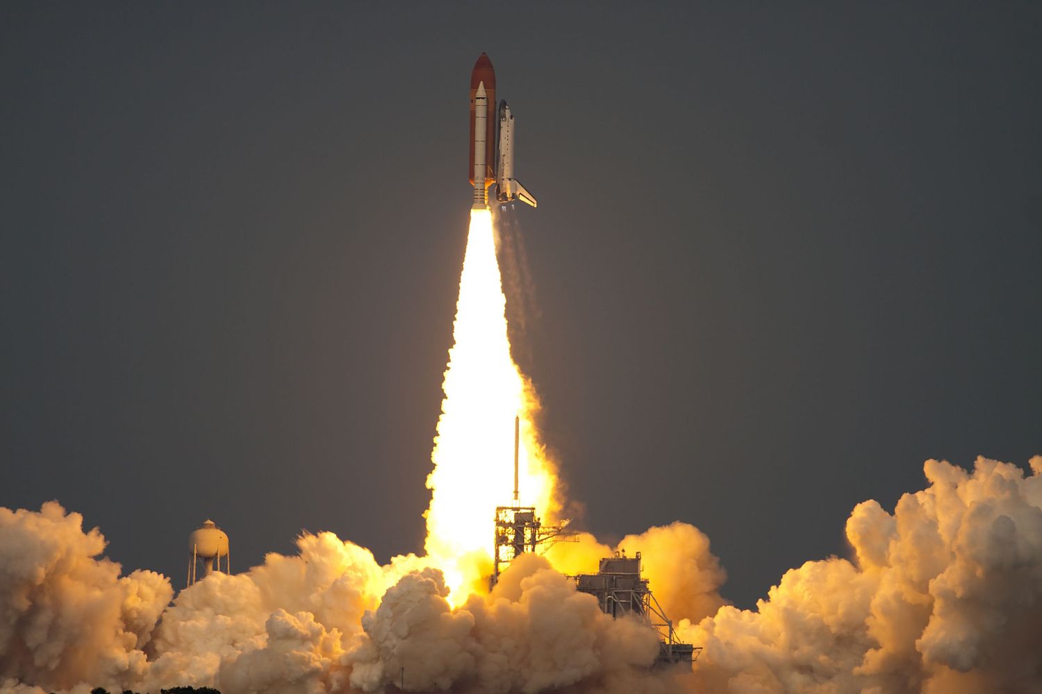 Space shuttle launch with bright flames and exhaust clouds against dark sky.