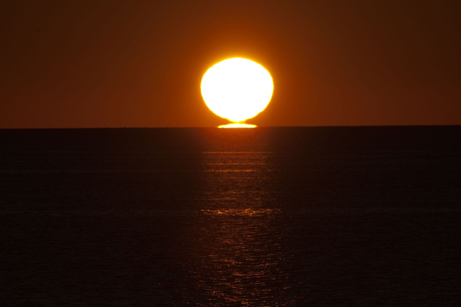 Large orange sun setting over dark ocean horizon creating dramatic reflection.