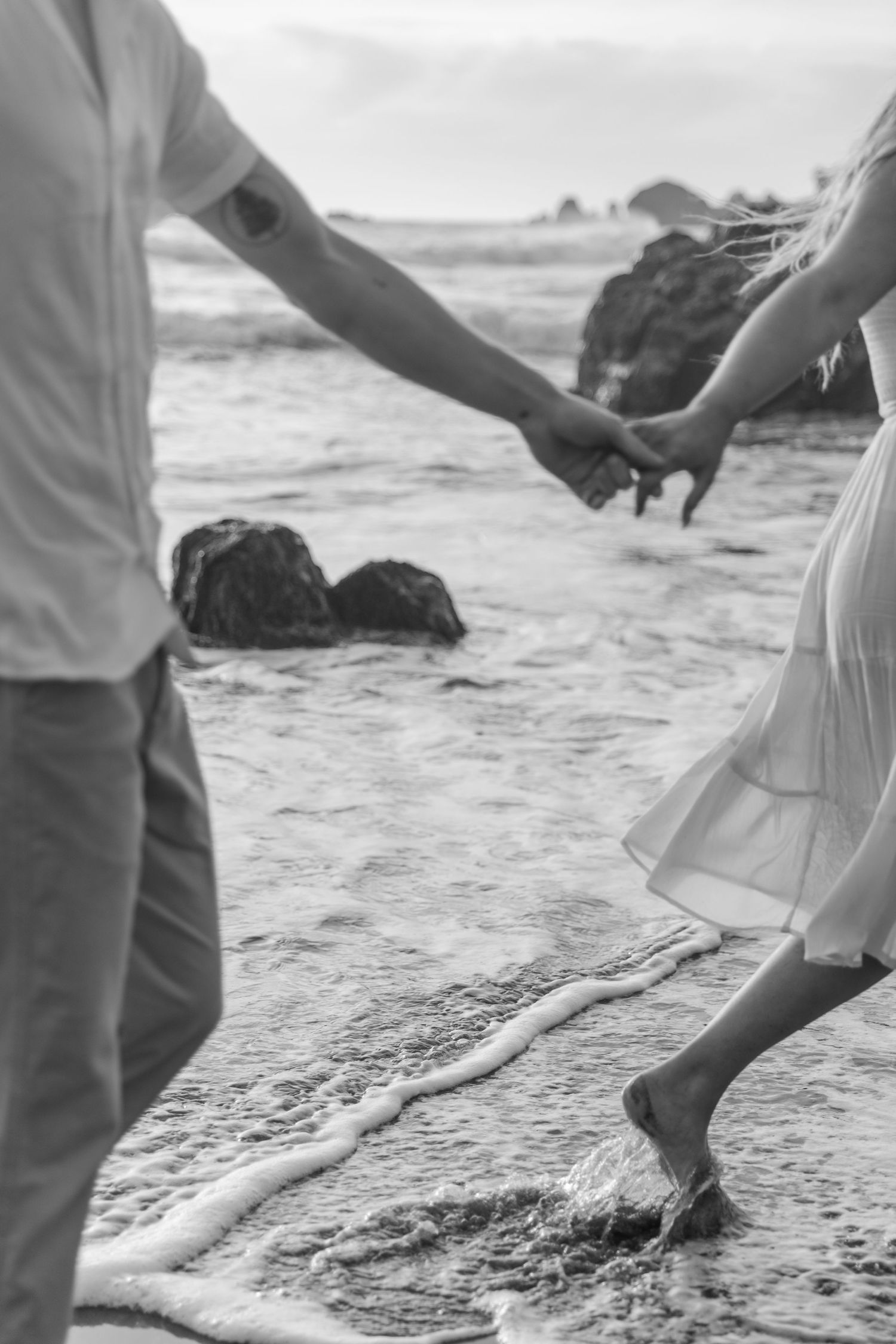 Black and white photo of feet walking along the shoreline as waves crash on a sandy beach.
