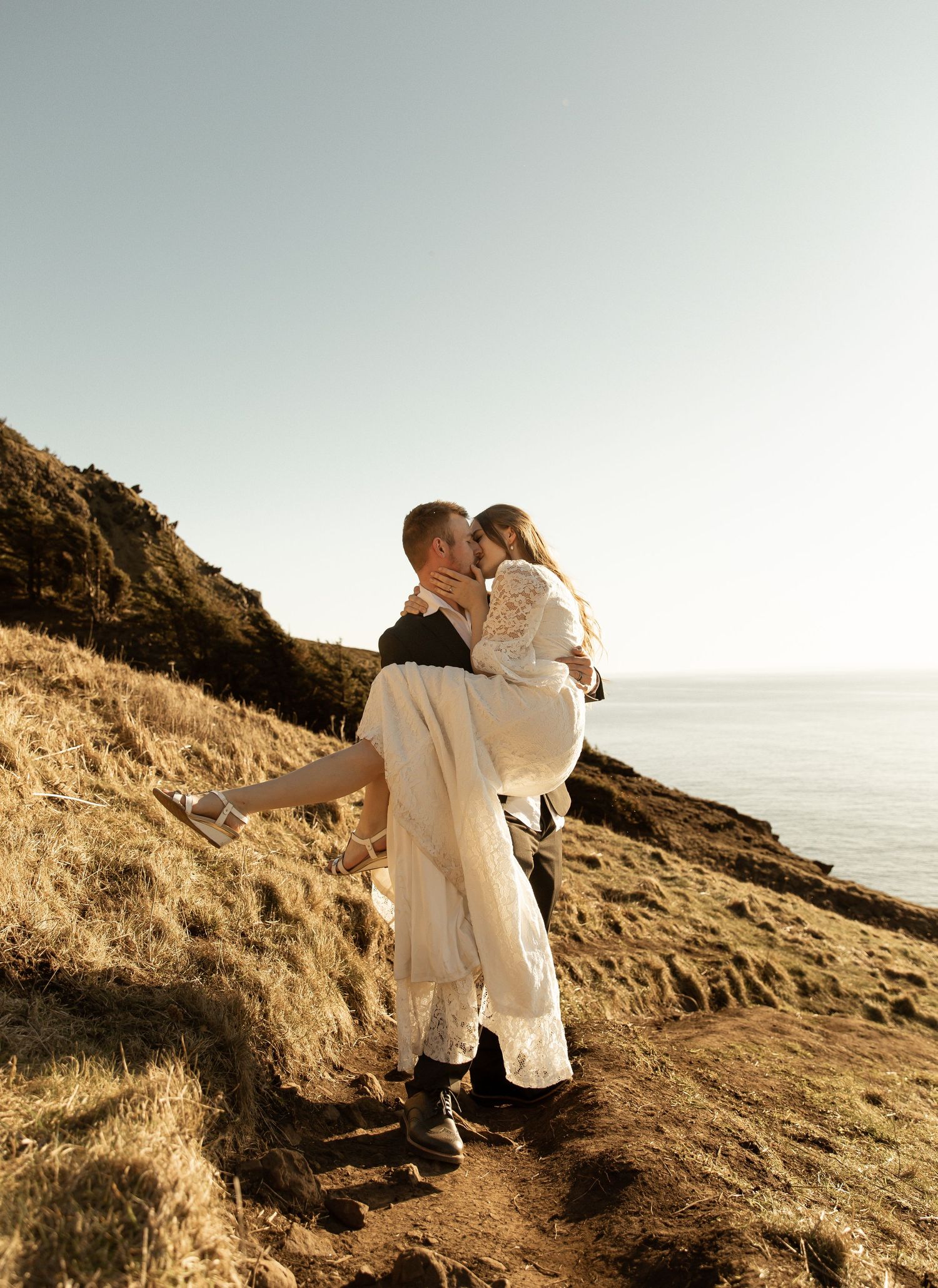 A couple shares a romantic moment on a coastal cliff overlooking the ocean at sunset.
