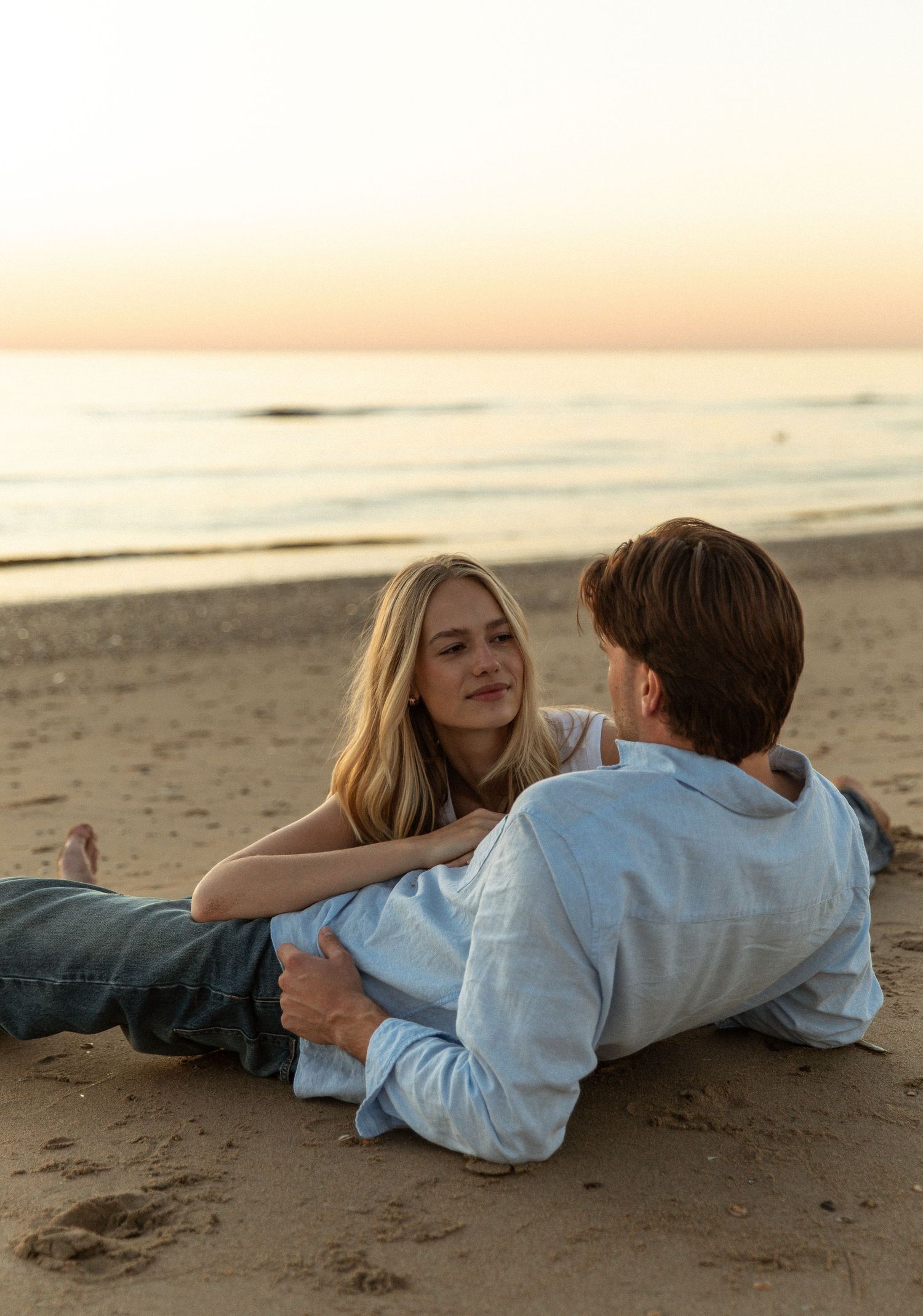 Couple lying on sandy beach at sunset, sharing a romantic moment with ocean waves in the background.