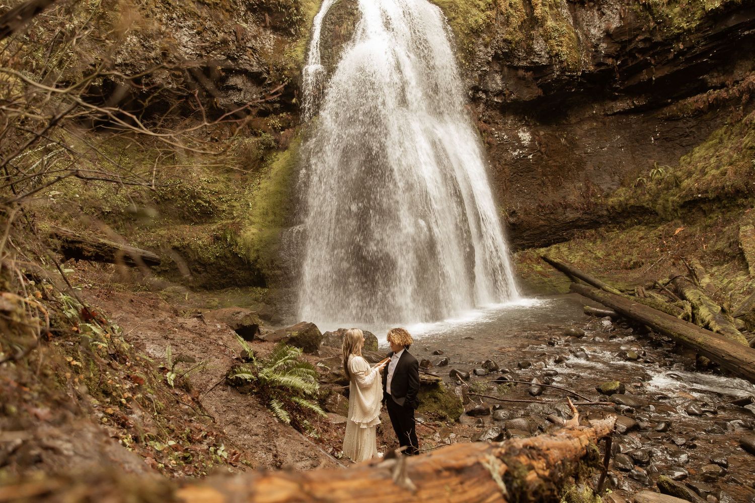 A dramatic waterfall flows over rocky cliffs while a couple poses in wedding attire at its base.
