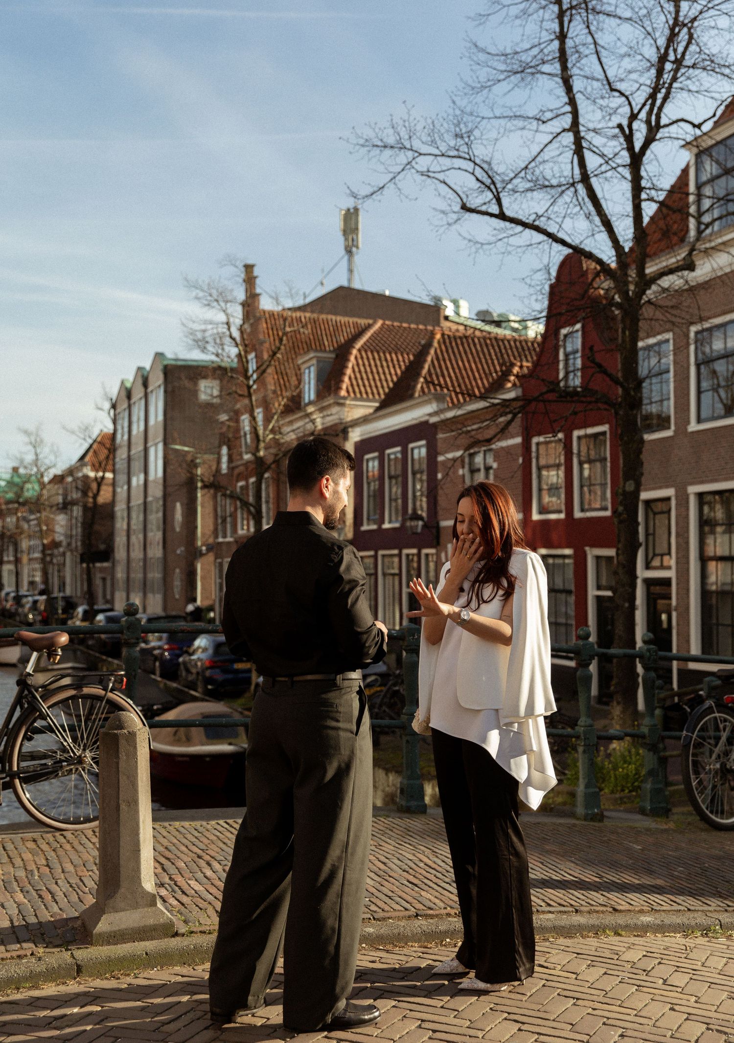 Couple sharing a romantic proposal moment on a cobblestone street in Amsterdam with historic canal houses in the background.