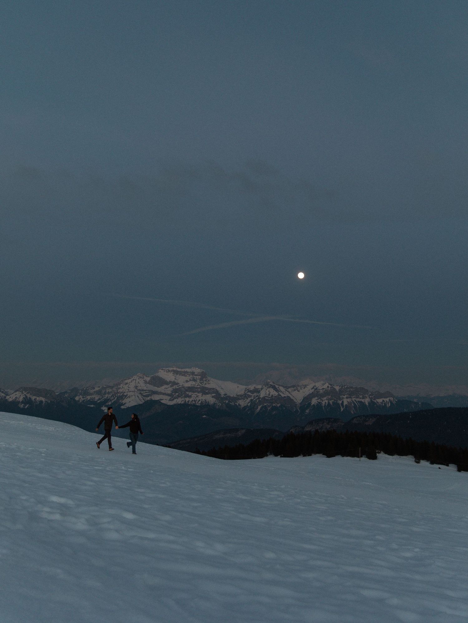 Skiers traverse a snowy mountain ridge at dusk with a full moon rising over snow-capped peaks in the distance.