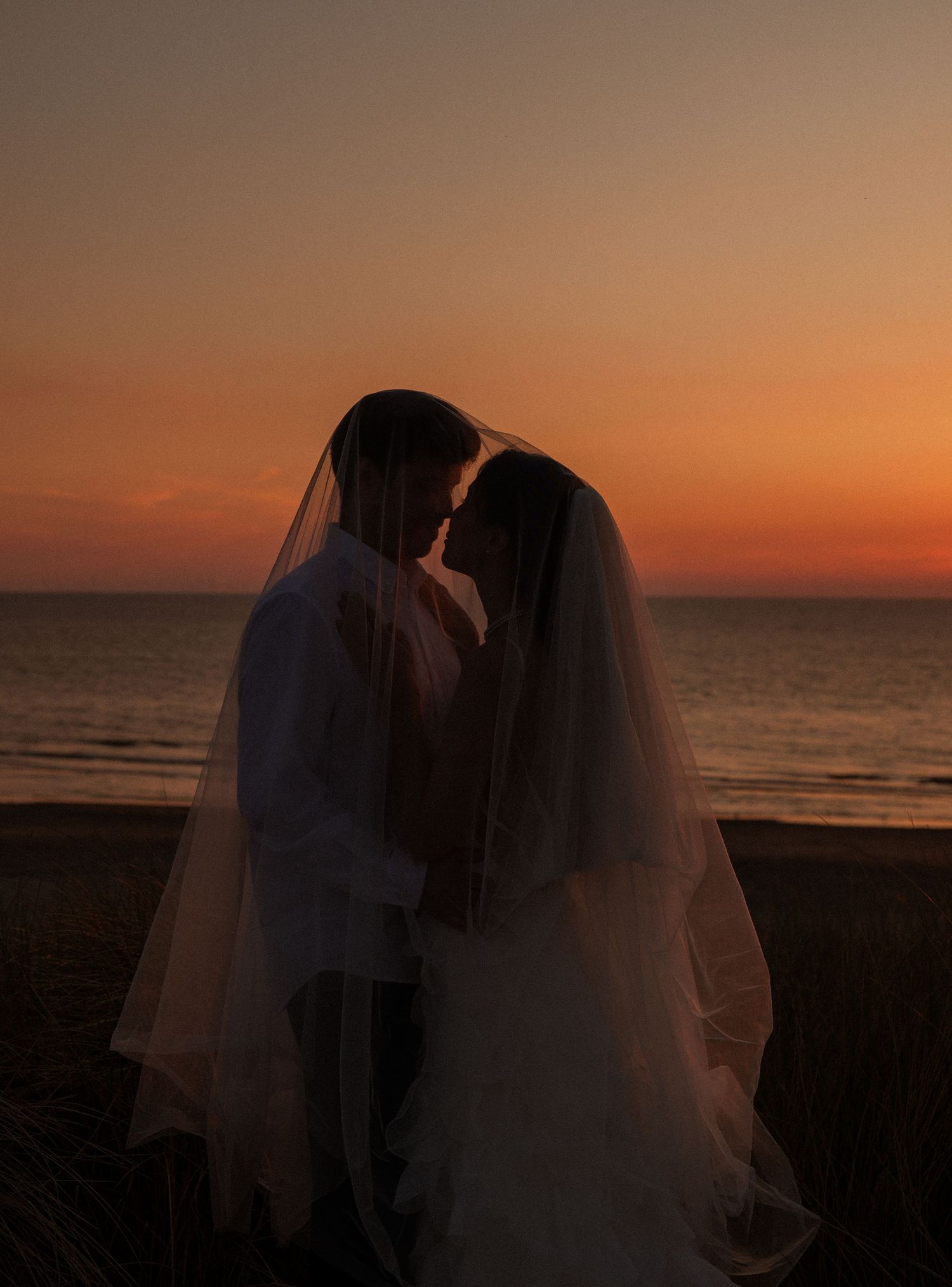 Romantic couple sharing a kiss in wedding attire silhouetted against a stunning orange sunset on the beach.