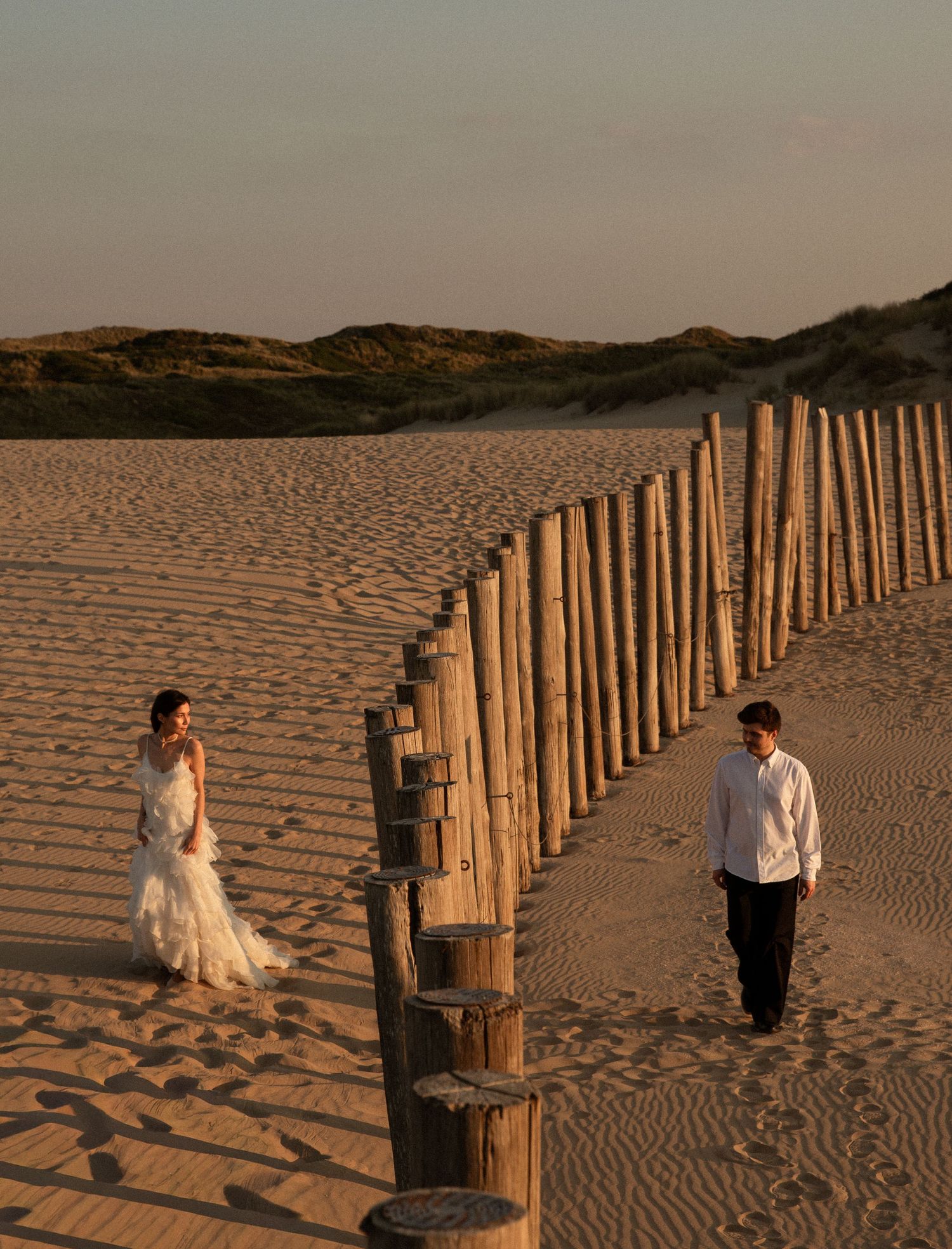 Bride and groom walk along a wooden fence on a sandy beach at golden hour sunset.