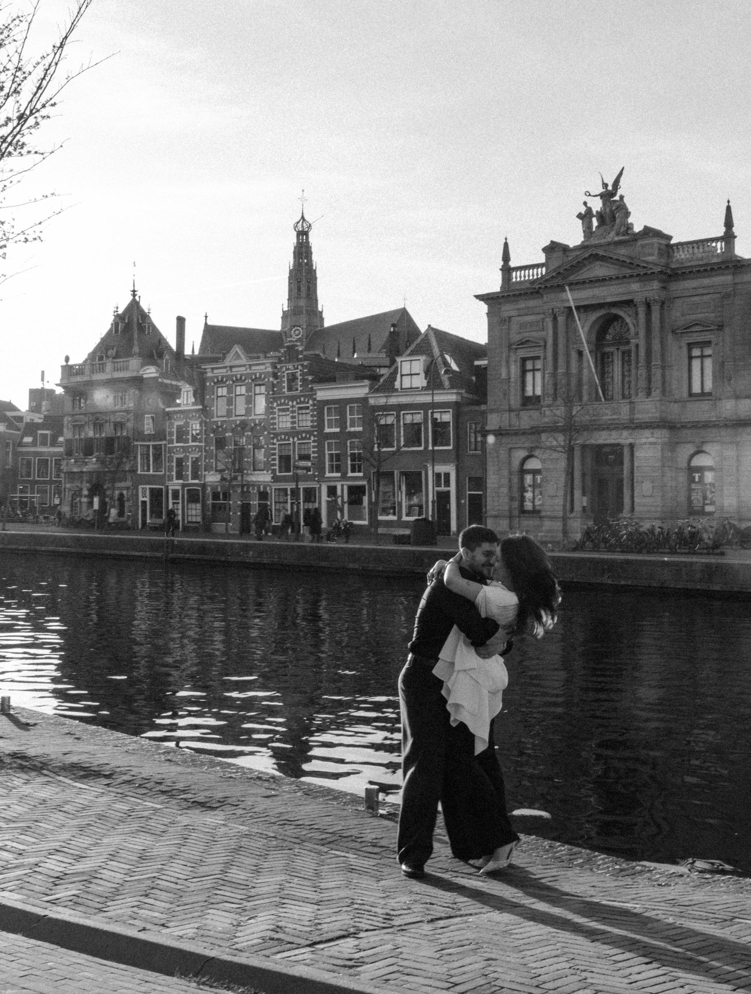 Black and white photo of a couple kissing by a canal in Haarlem, Netherlands, with historic buildings in the background.