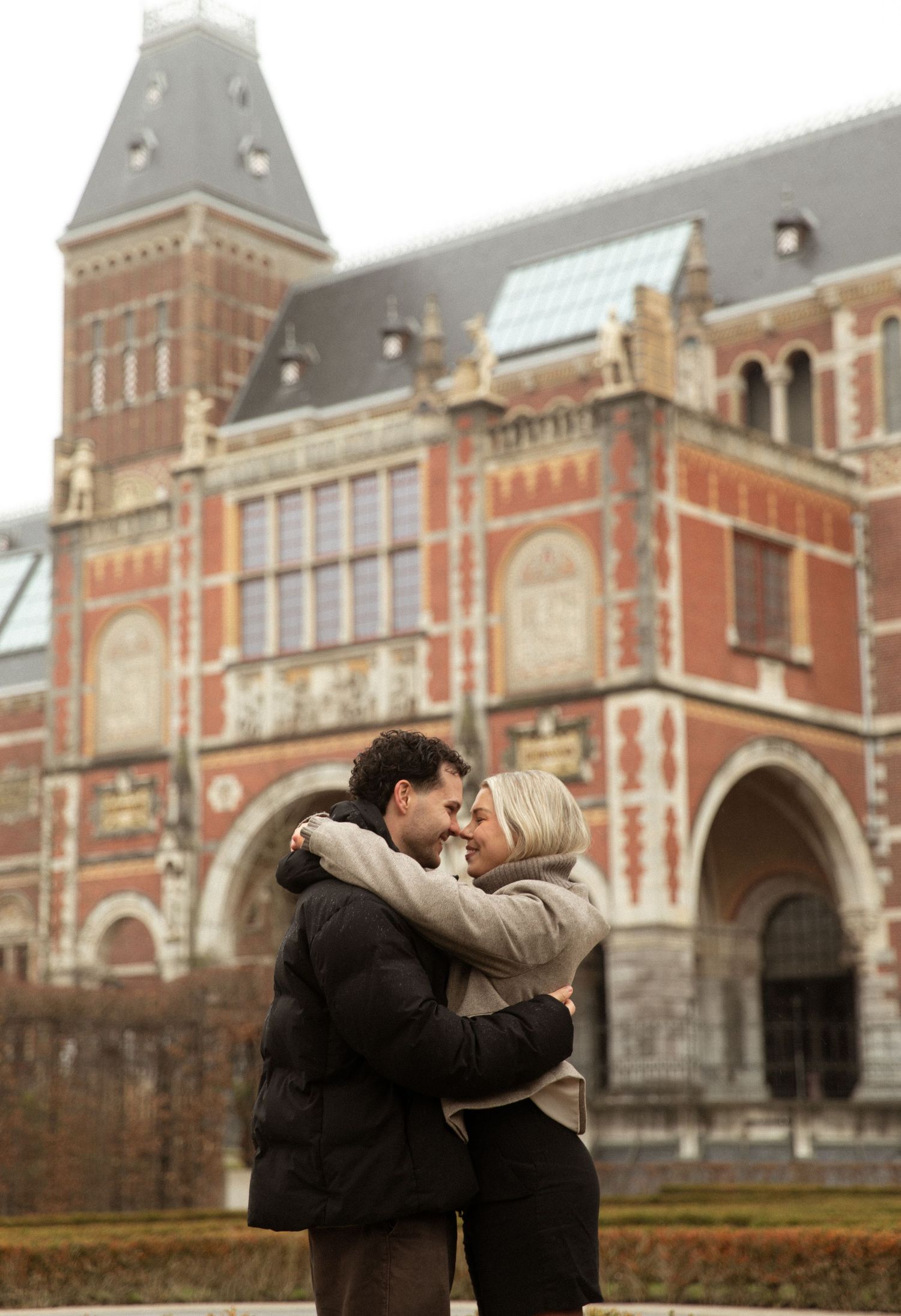 A romantic couple embraces in front of the historic Rijksmuseum building in Amsterdam on an overcast day.