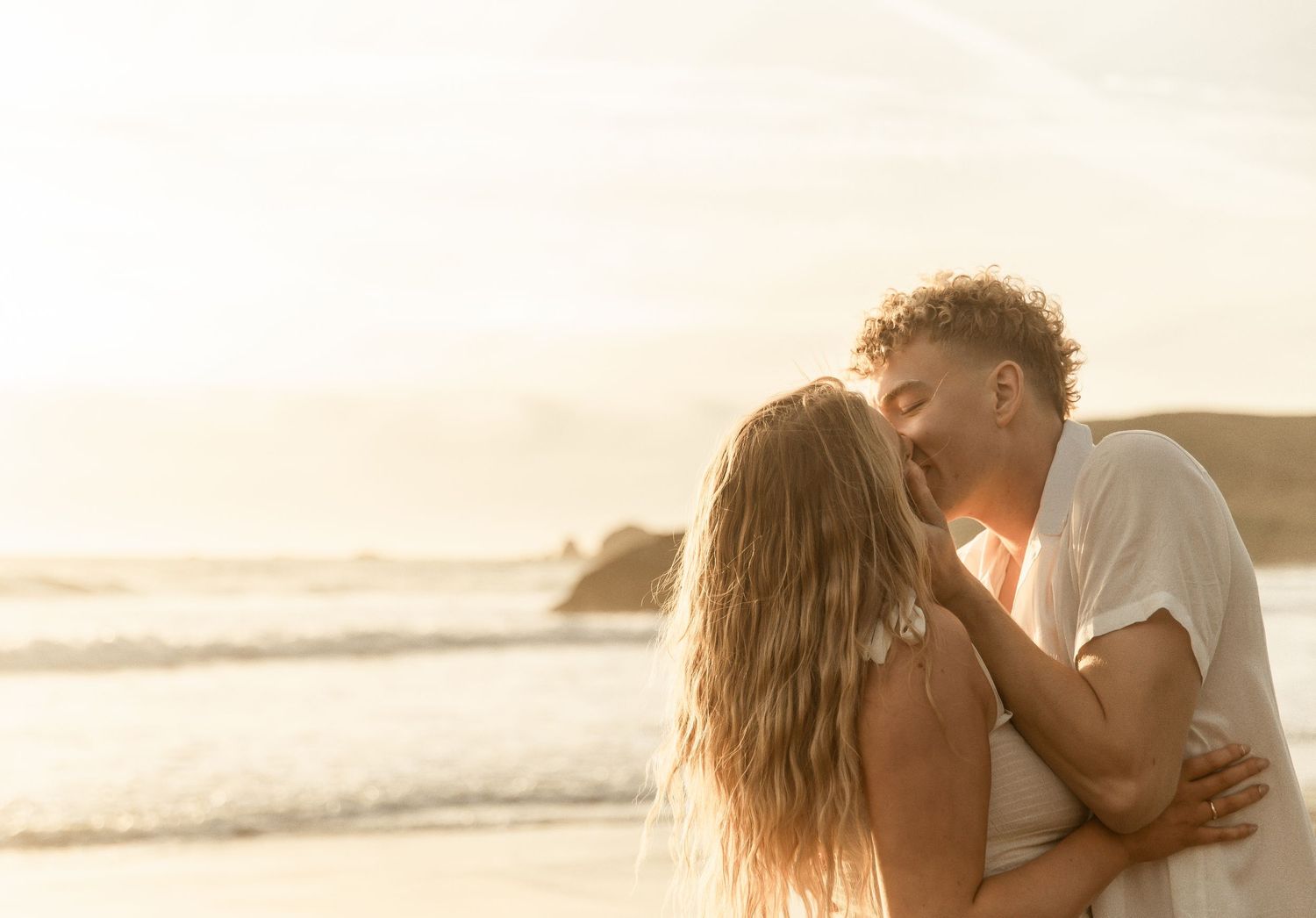 A romantic couple shares an intimate moment at sunset on a sandy beach during their engagement photoshoot.