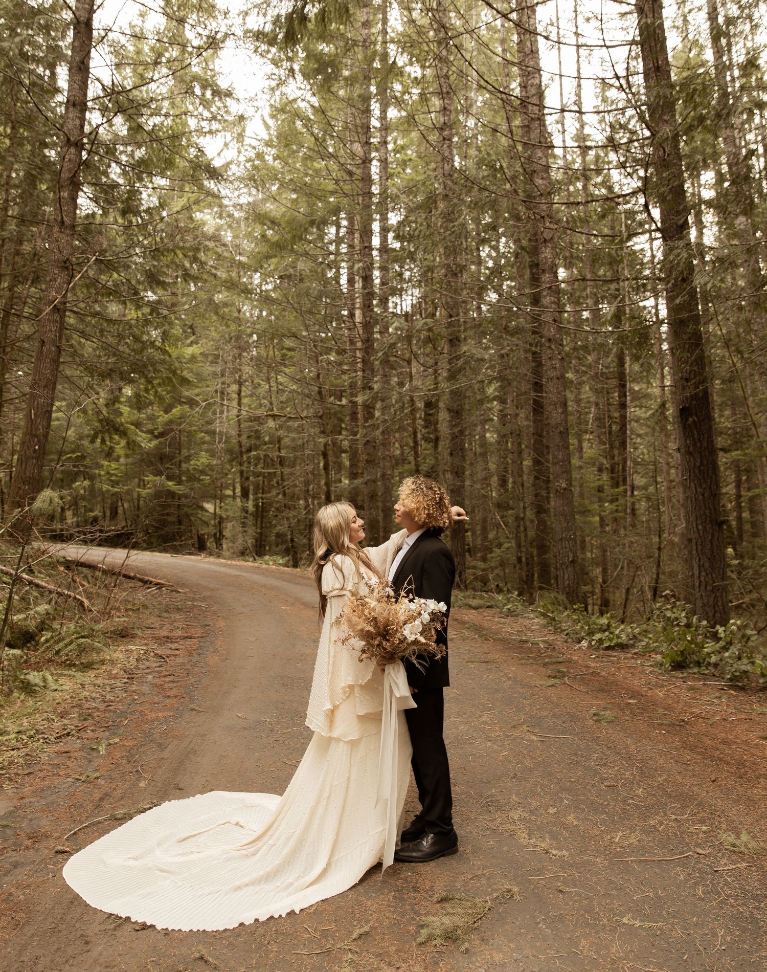 A romantic forest wedding photoshoot with a flowing white dress and black suit on a winding dirt path.