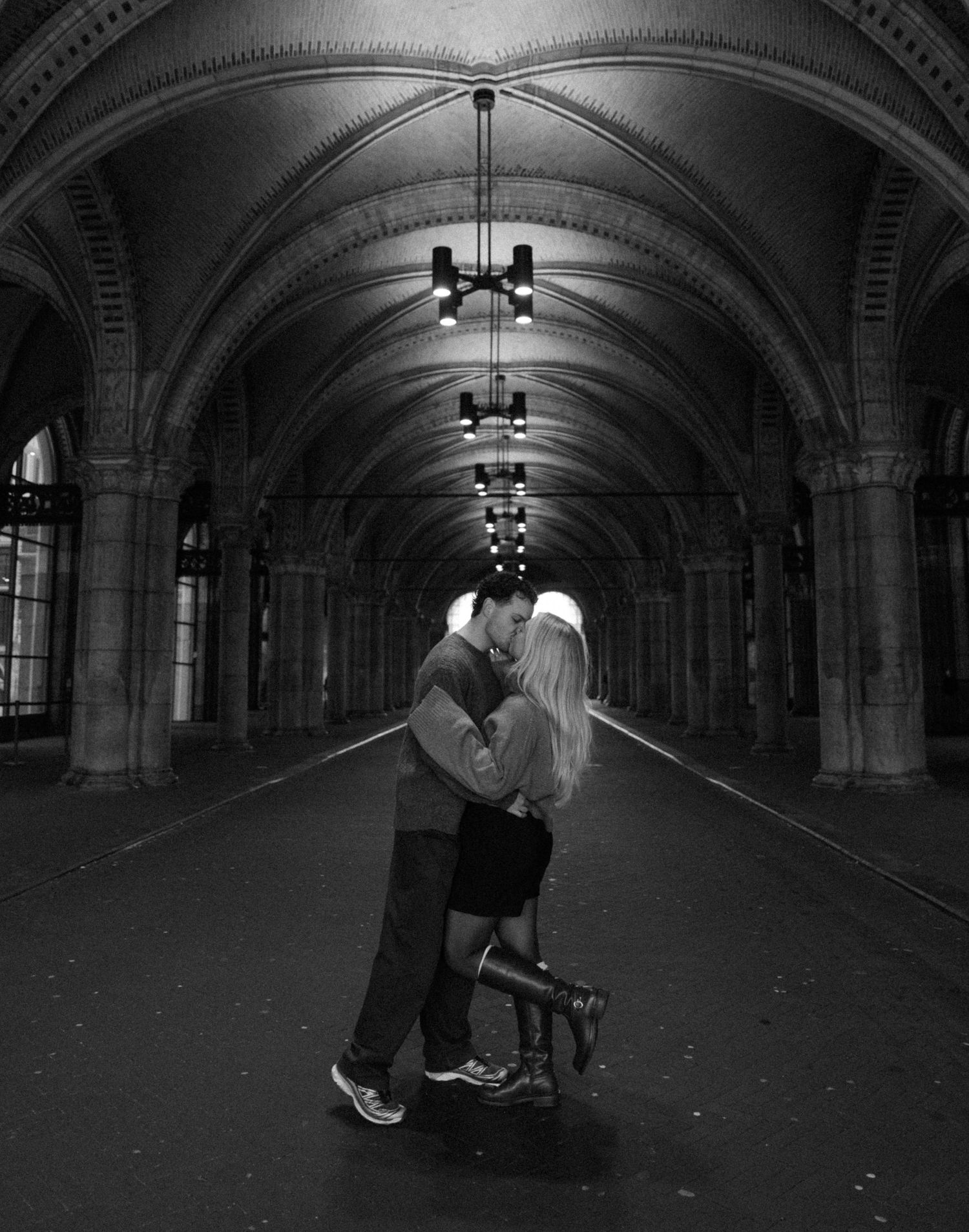 Black and white photo of couple kissing in dramatic arched corridor with hanging lights.