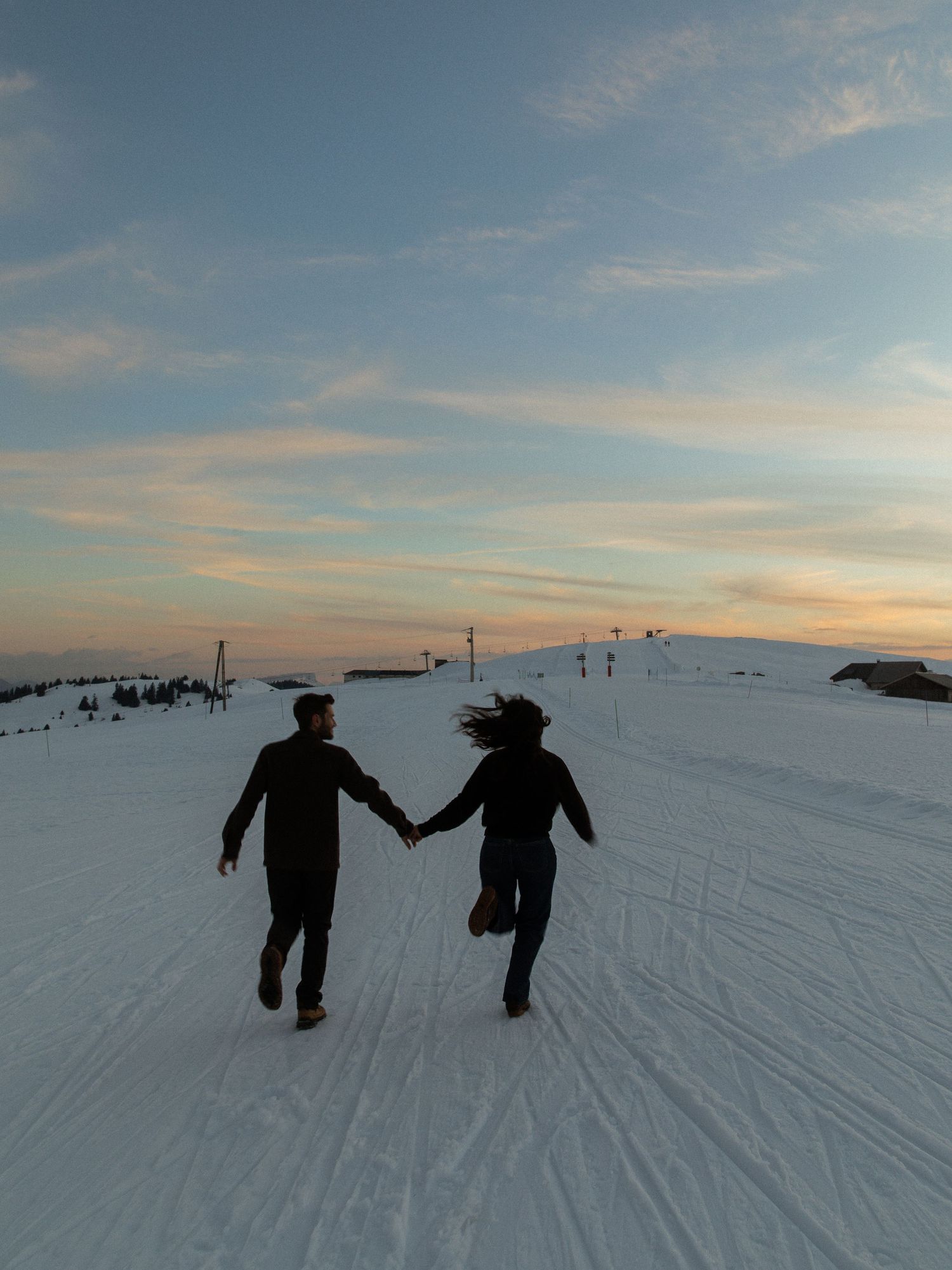 Two silhouettes walk hand in hand across a snowy landscape at sunset with ski lifts visible in the distance.