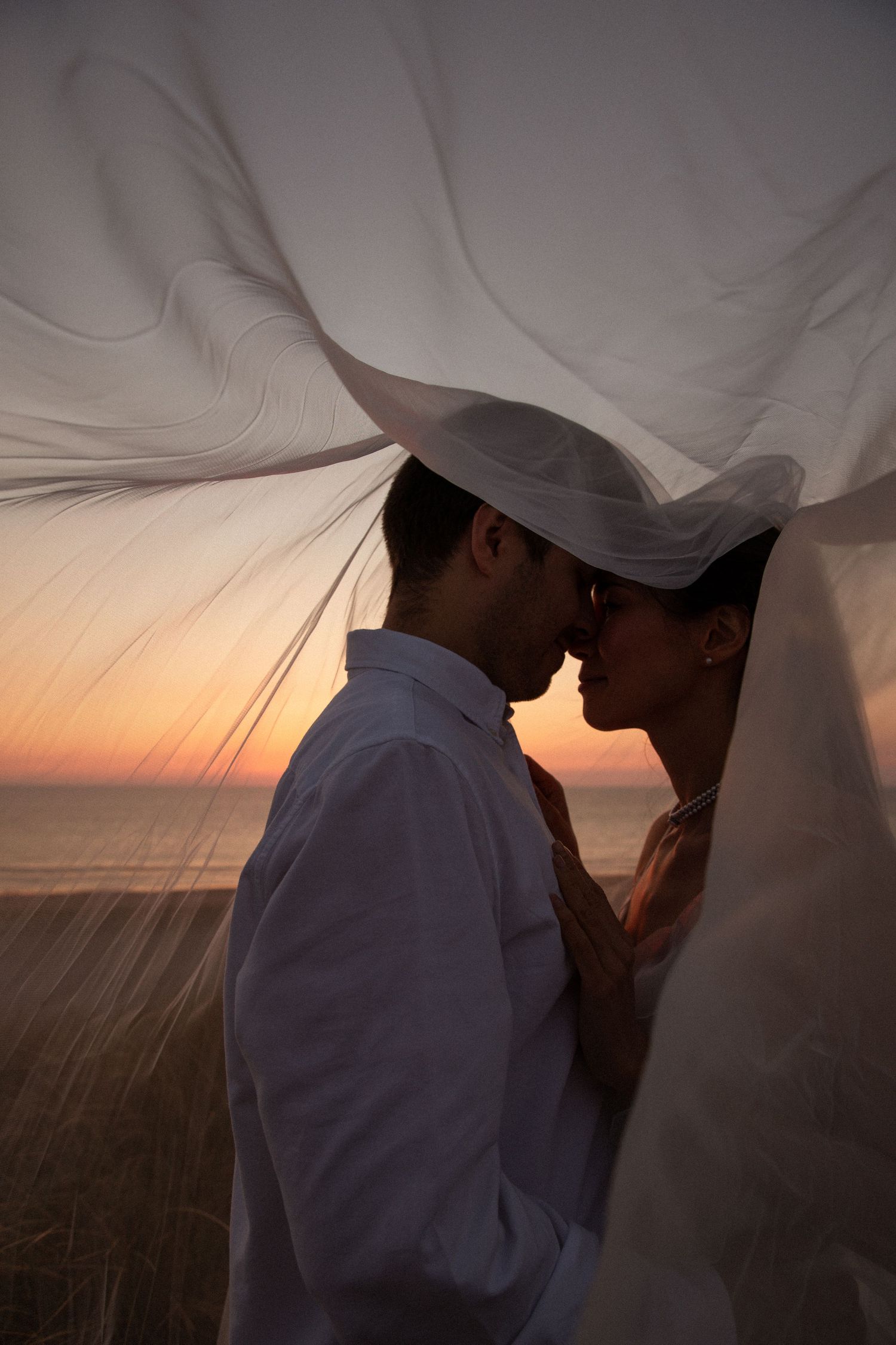 Couple sharing a romantic moment under a flowing white veil at sunset by the ocean.