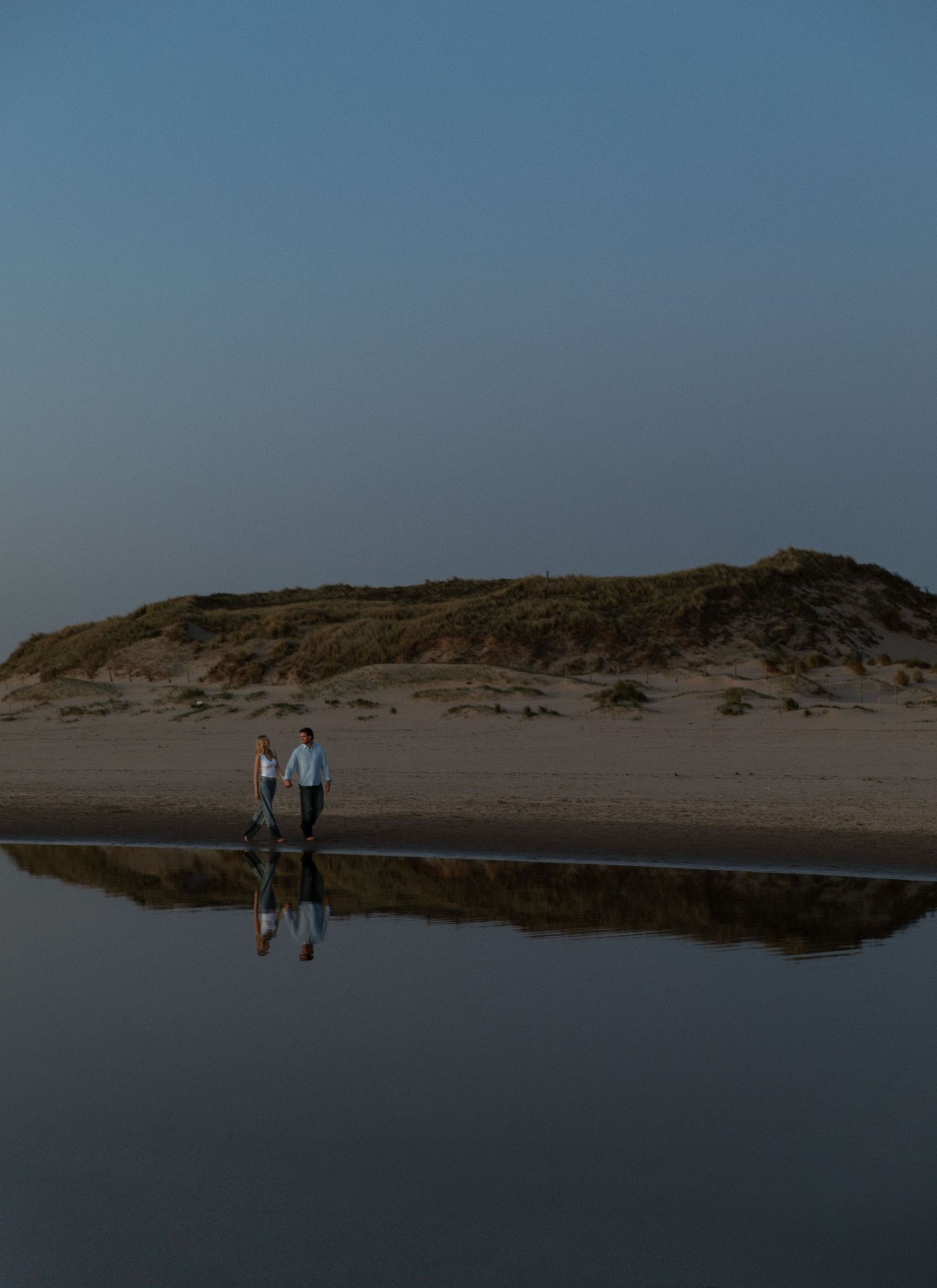 Couple holding hands on a sandy beach at dusk, reflected in still water, with sand dunes in the background.