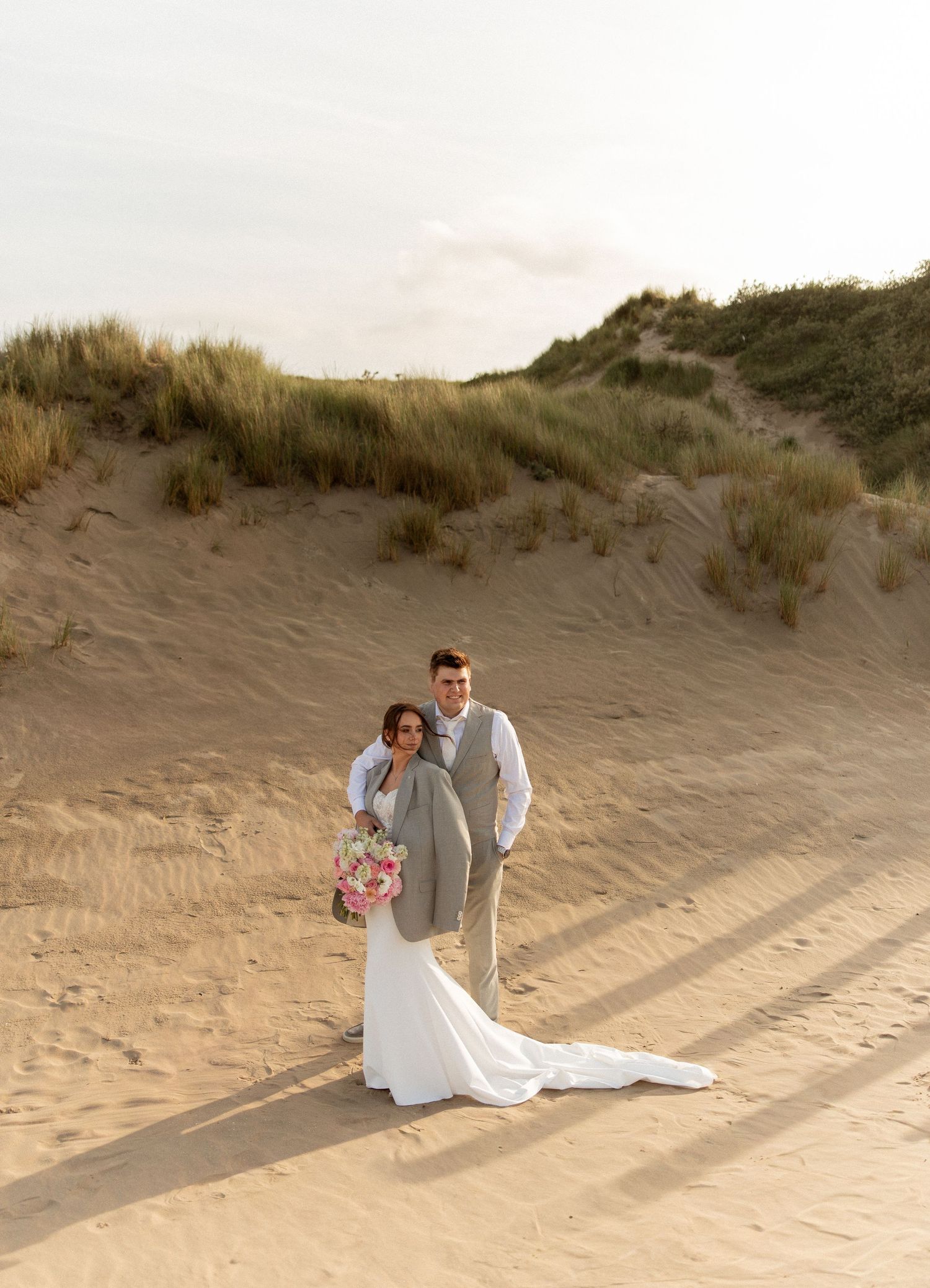 Bride and groom posing on sandy beach dunes at golden hour, bride holding pink bouquet in elegant white gown.