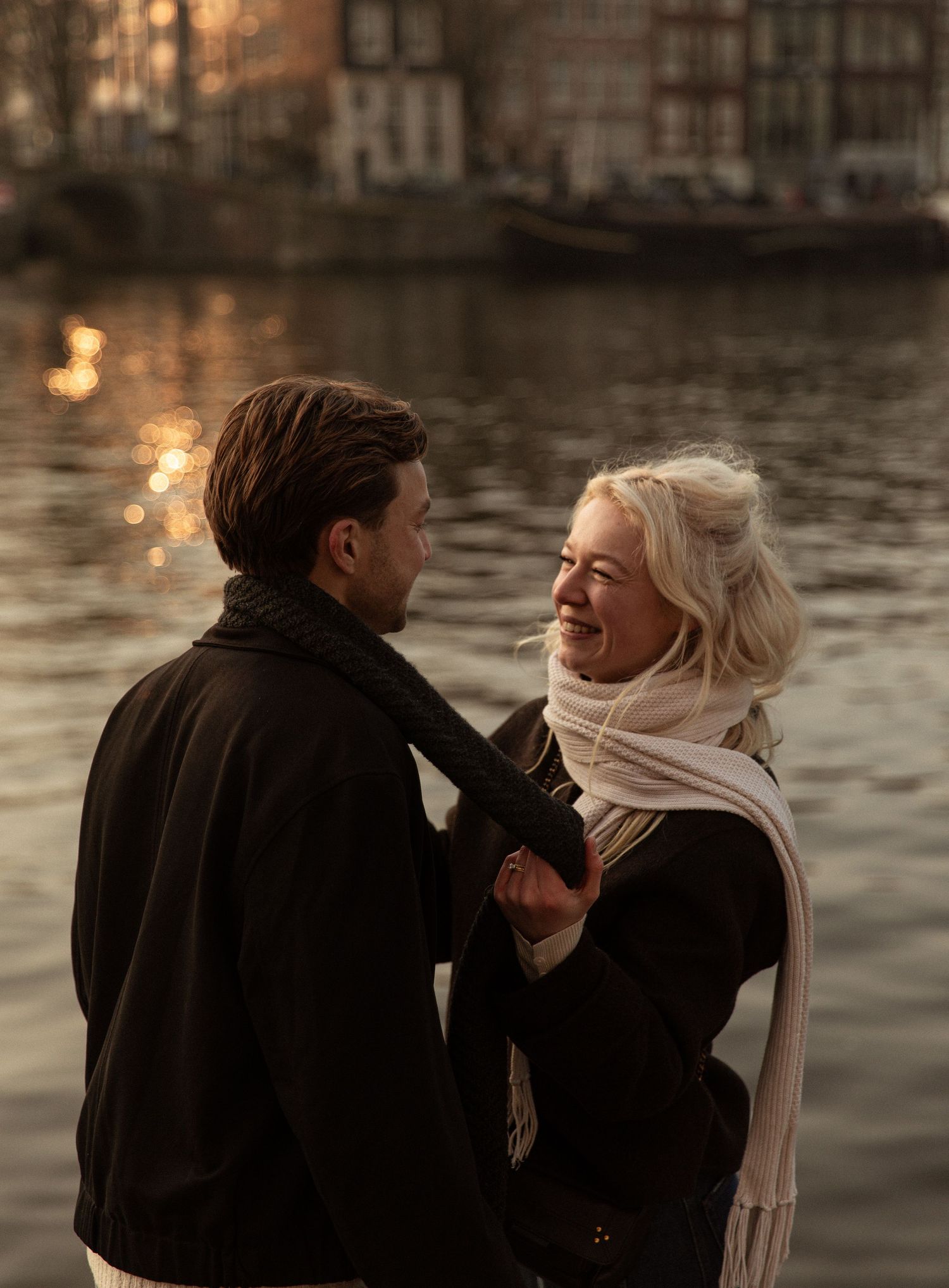 Couple smiling at each other by a sunlit canal in Amsterdam, woman in white scarf, golden hour backdrop.