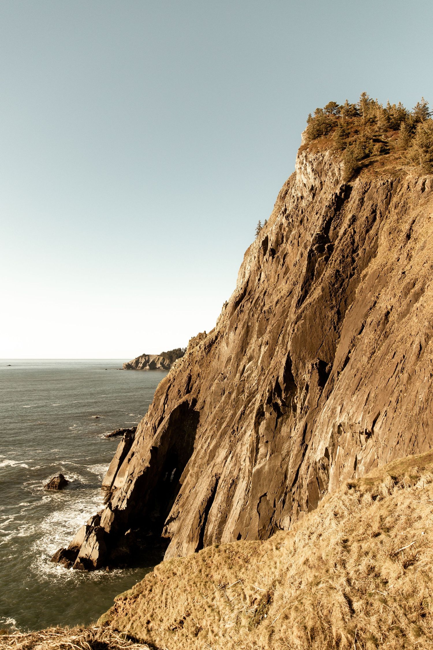 Dramatic coastal cliffs rise above the Pacific Ocean with rugged rock formations and waves crashing below.