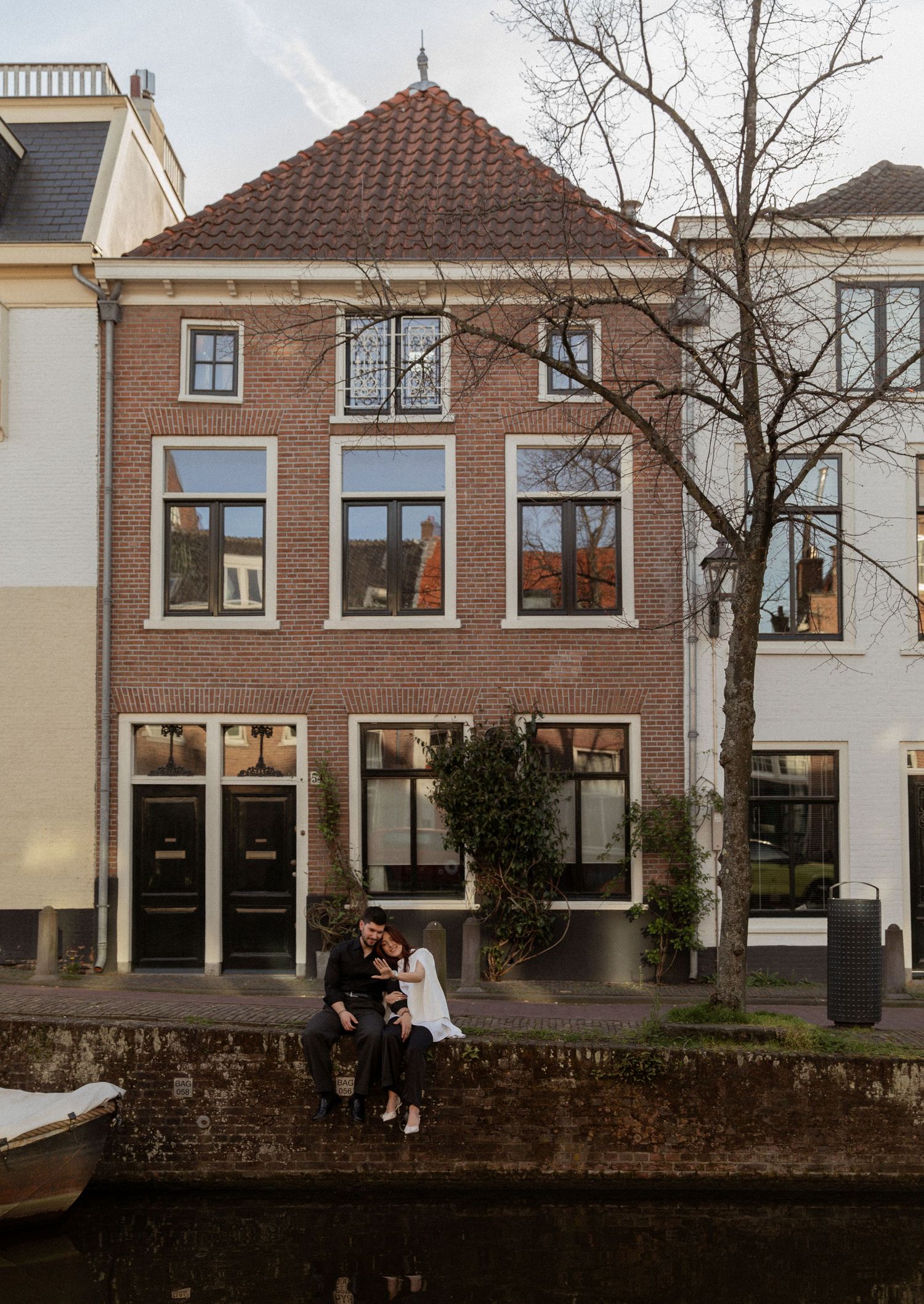 Couple sitting on a canal wall in front of a charming Dutch brick townhouse with red tile roof.