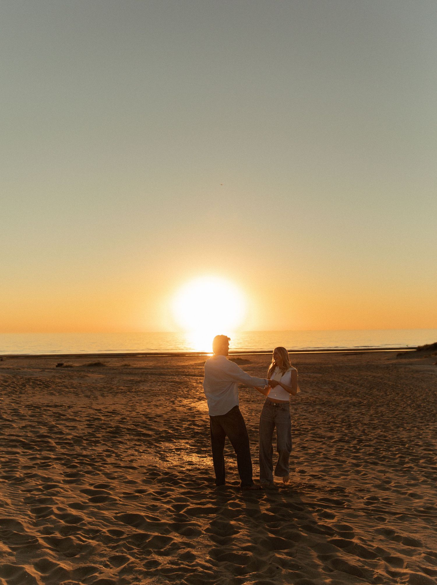 Couple holding hands on sandy beach at golden sunset with sun setting over calm ocean horizon.