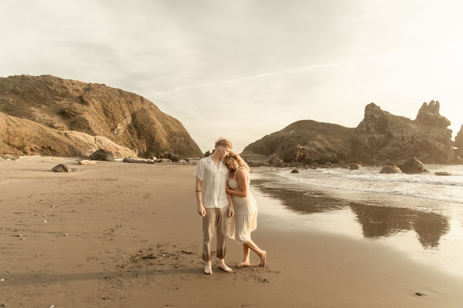 A couple in white clothing walks their dog along a scenic coastal beach with dramatic cliffs and cloudy skies.