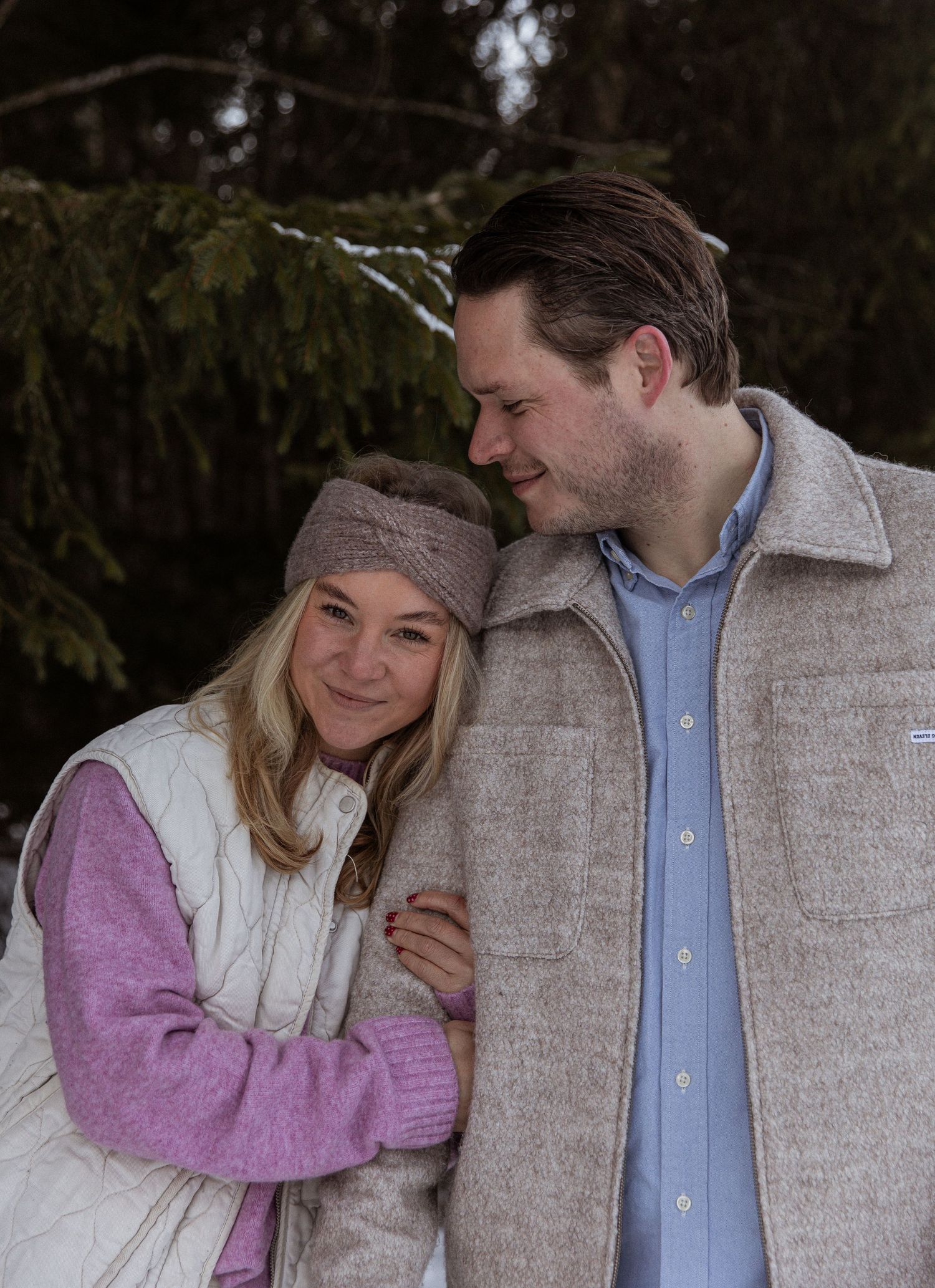 A cozy winter portrait of a couple in warm clothing against an evergreen backdrop.