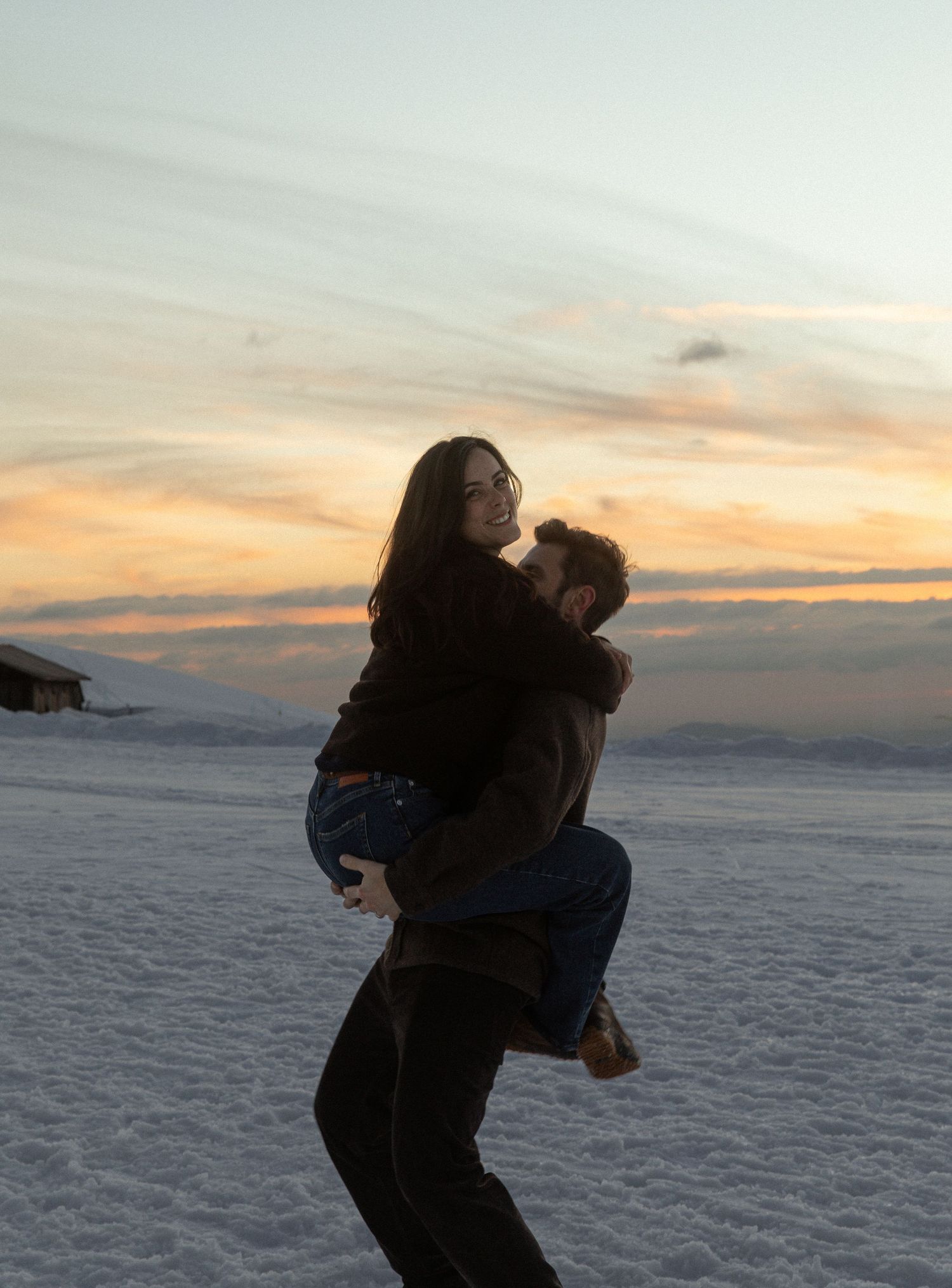 Two figures playfully dance together against a snowy winter landscape at sunset with dramatic orange sky.
