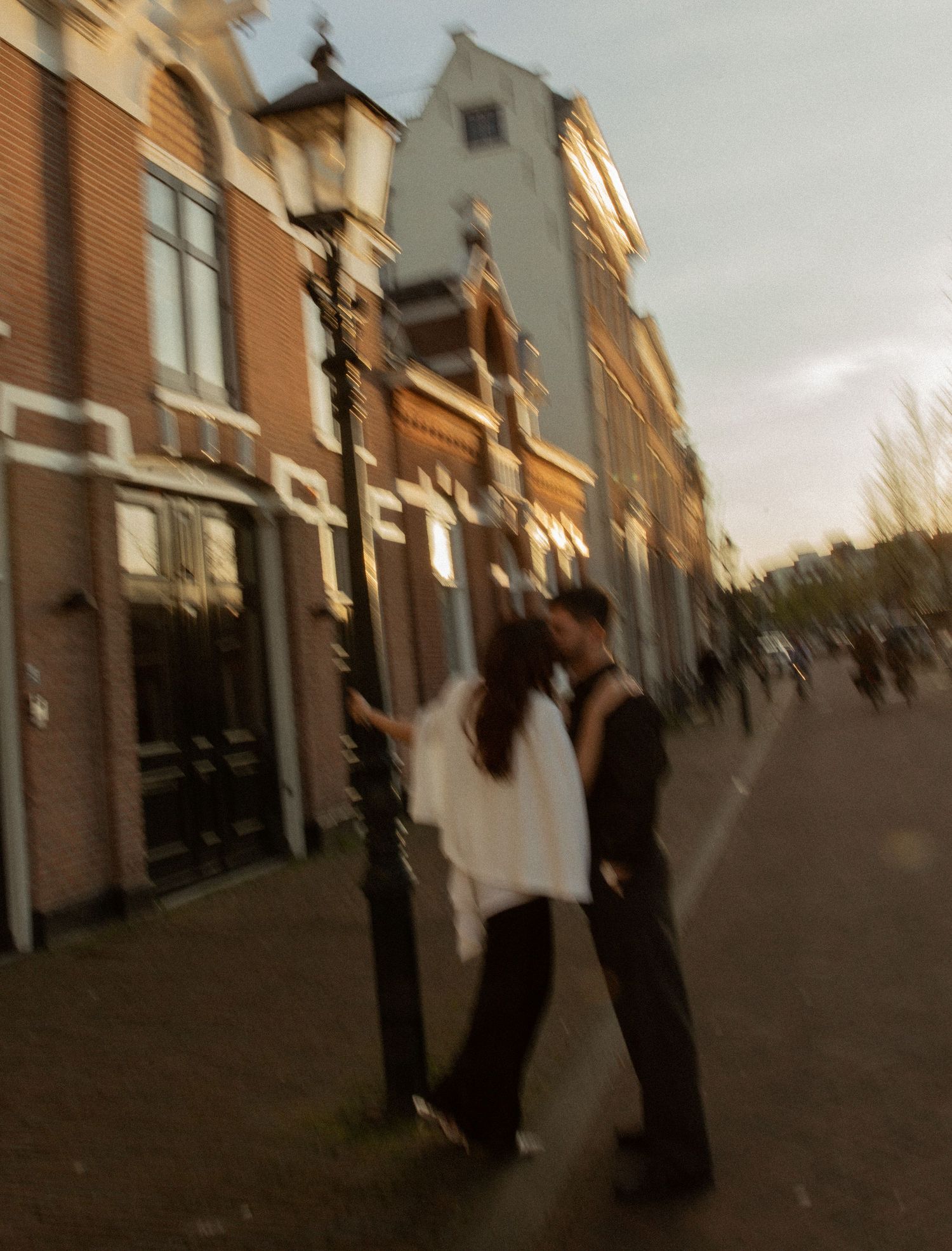 A couple shares a romantic kiss by a vintage street lamp on a cobblestone street in Amsterdam at dusk.