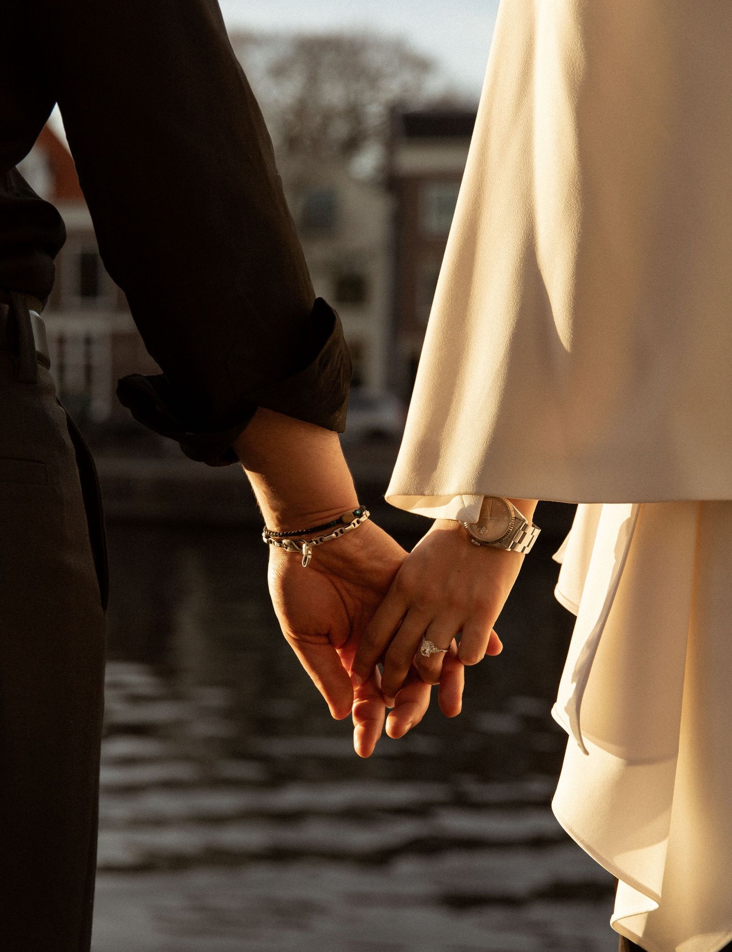 Couple holding hands in wedding attire, bride in white gown with bracelet and ring, groom in dark suit near waterfront.