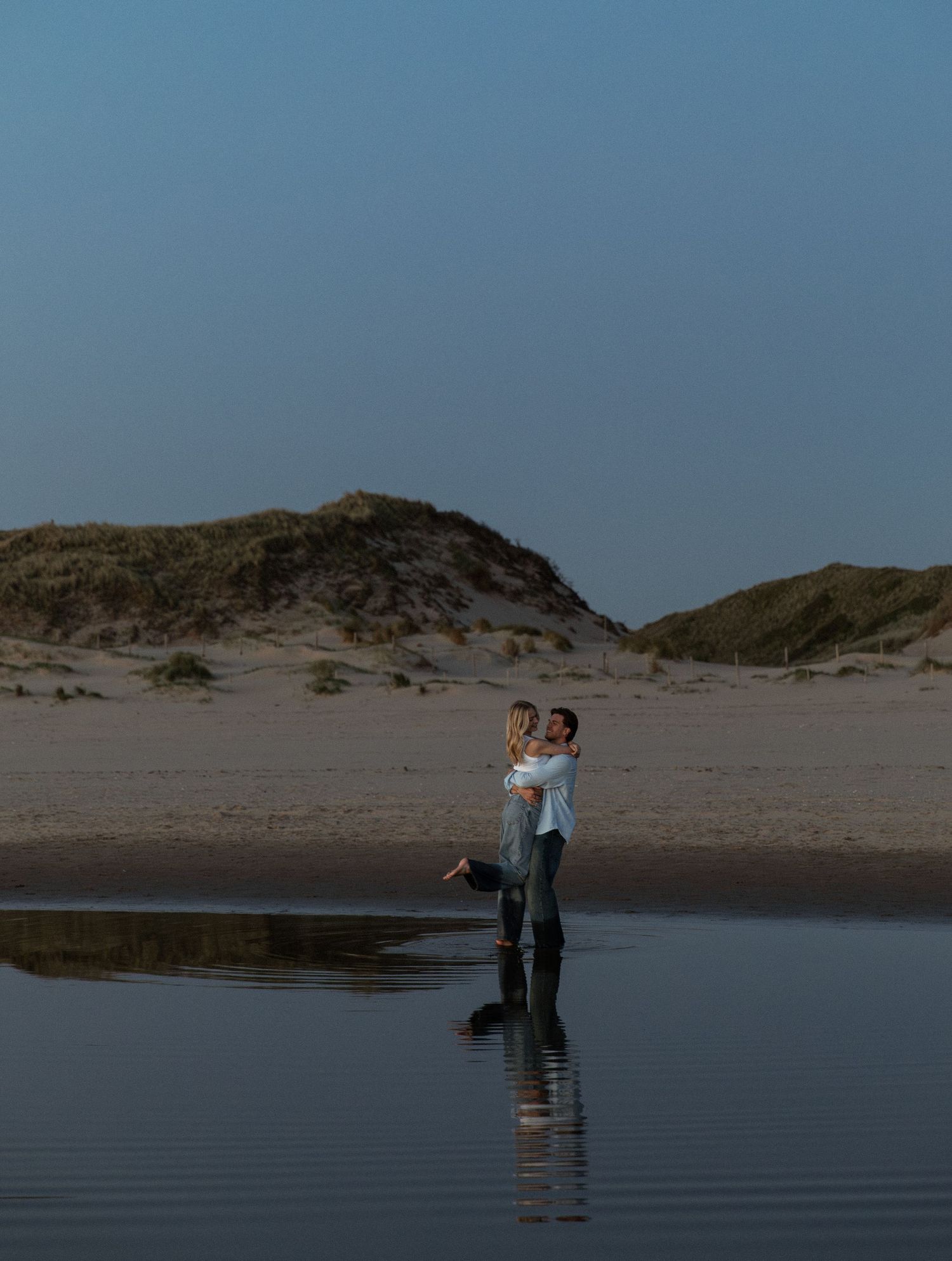 Couple embracing on a reflective beach at dusk with sand dunes in the background.