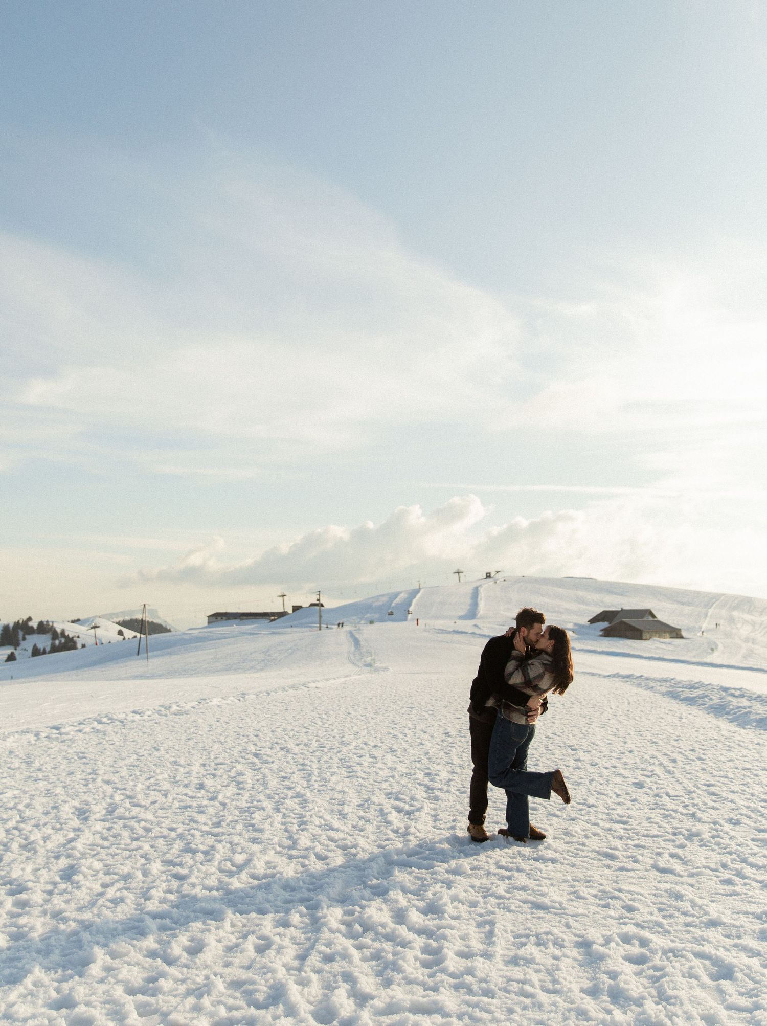 A couple stands together on a snow-covered mountain summit with a bright winter sky and white landscape in the background.