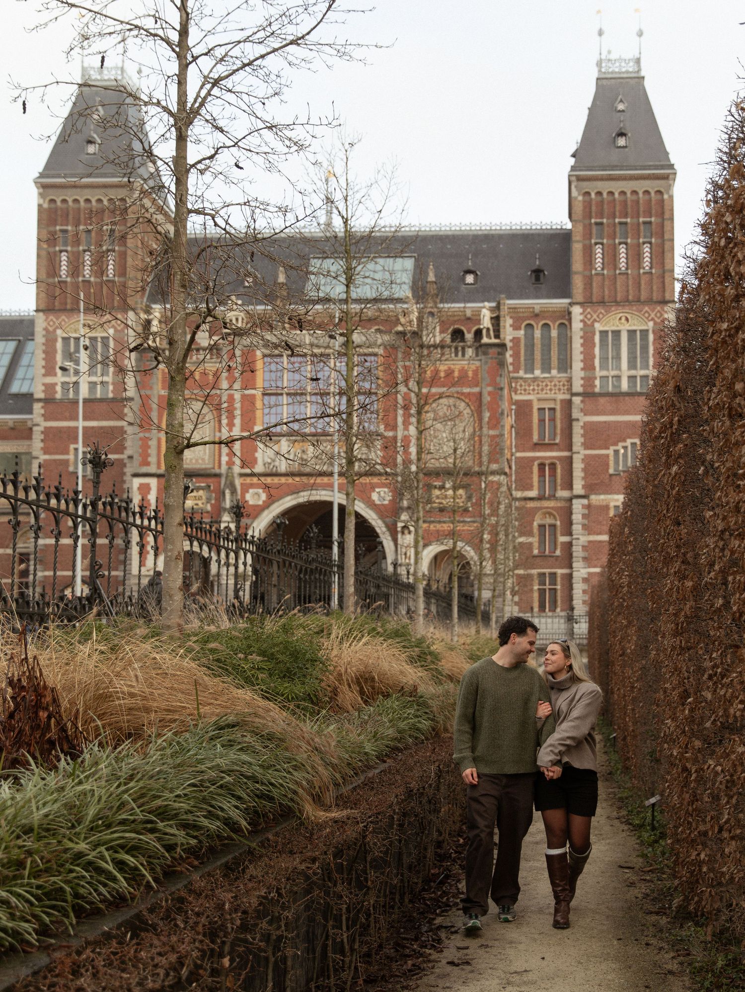 A couple strolls along a garden path in front of the historic Rijksmuseum building in Amsterdam on an autumn day.