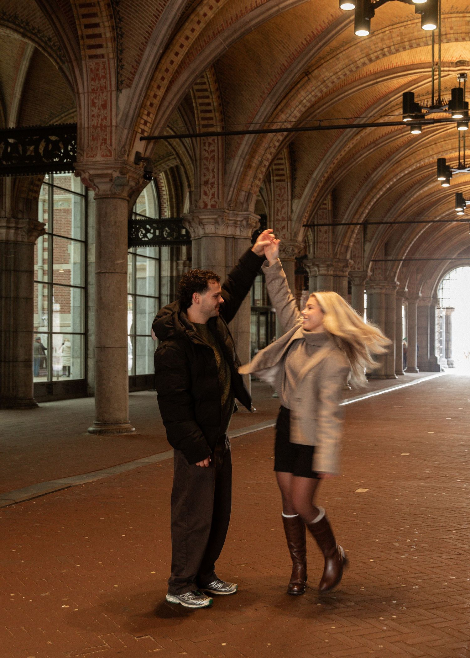 Two people dance together in an elegant arched hallway with ornate architectural details and warm lighting.