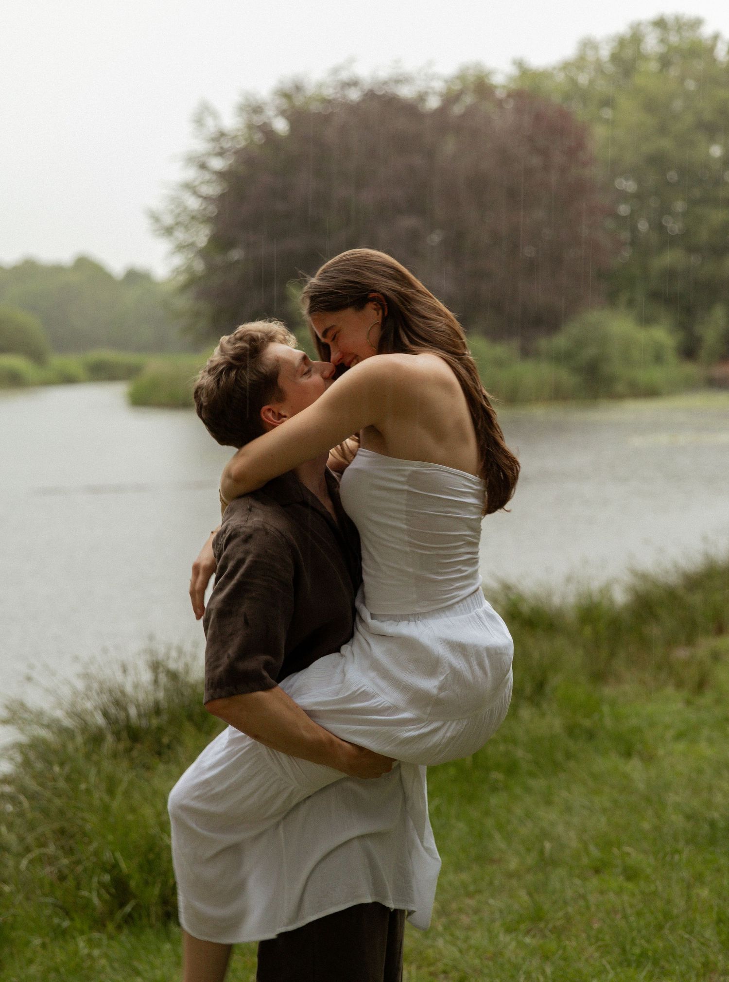 Romantic couple embracing and kissing in the rain near a serene lake surrounded by lush green trees.