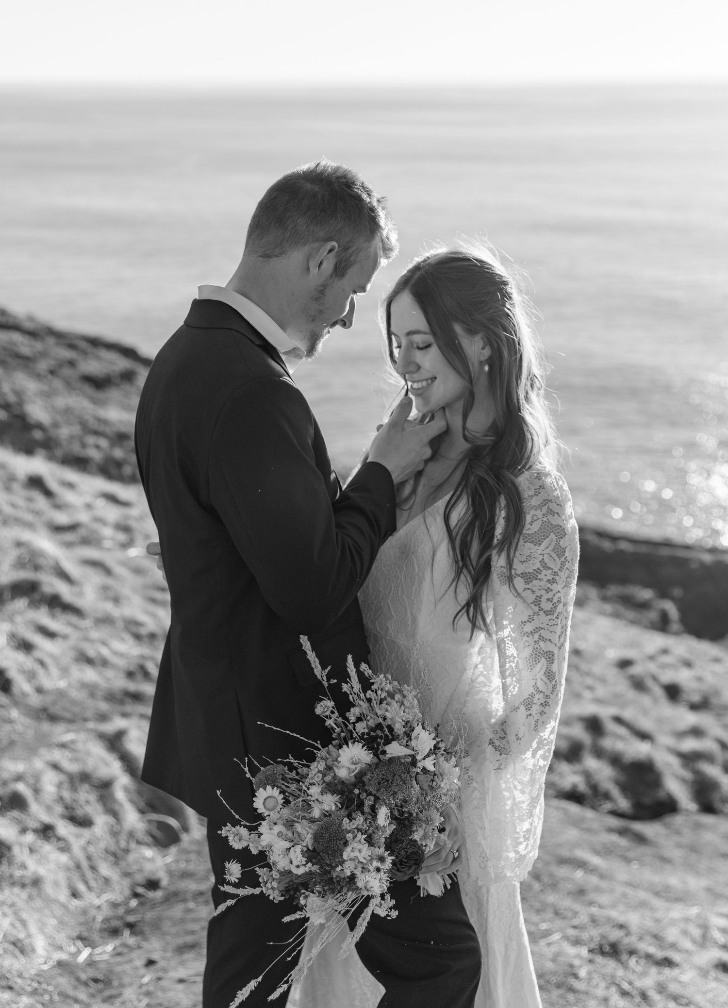 Black and white coastal wedding photos show a romantic couple embracing on a scenic cliff overlooking the ocean.