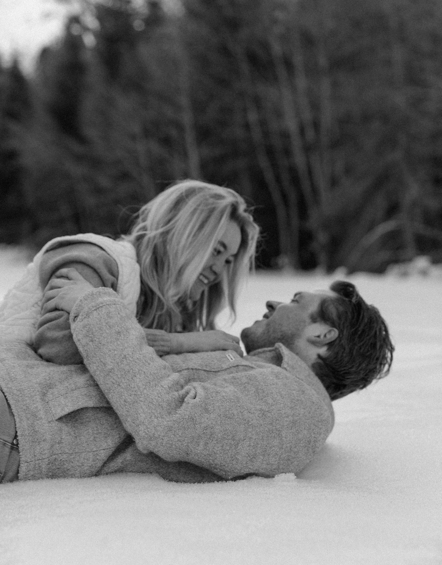 A playful black and white moment captured of two people lying in the snow together.