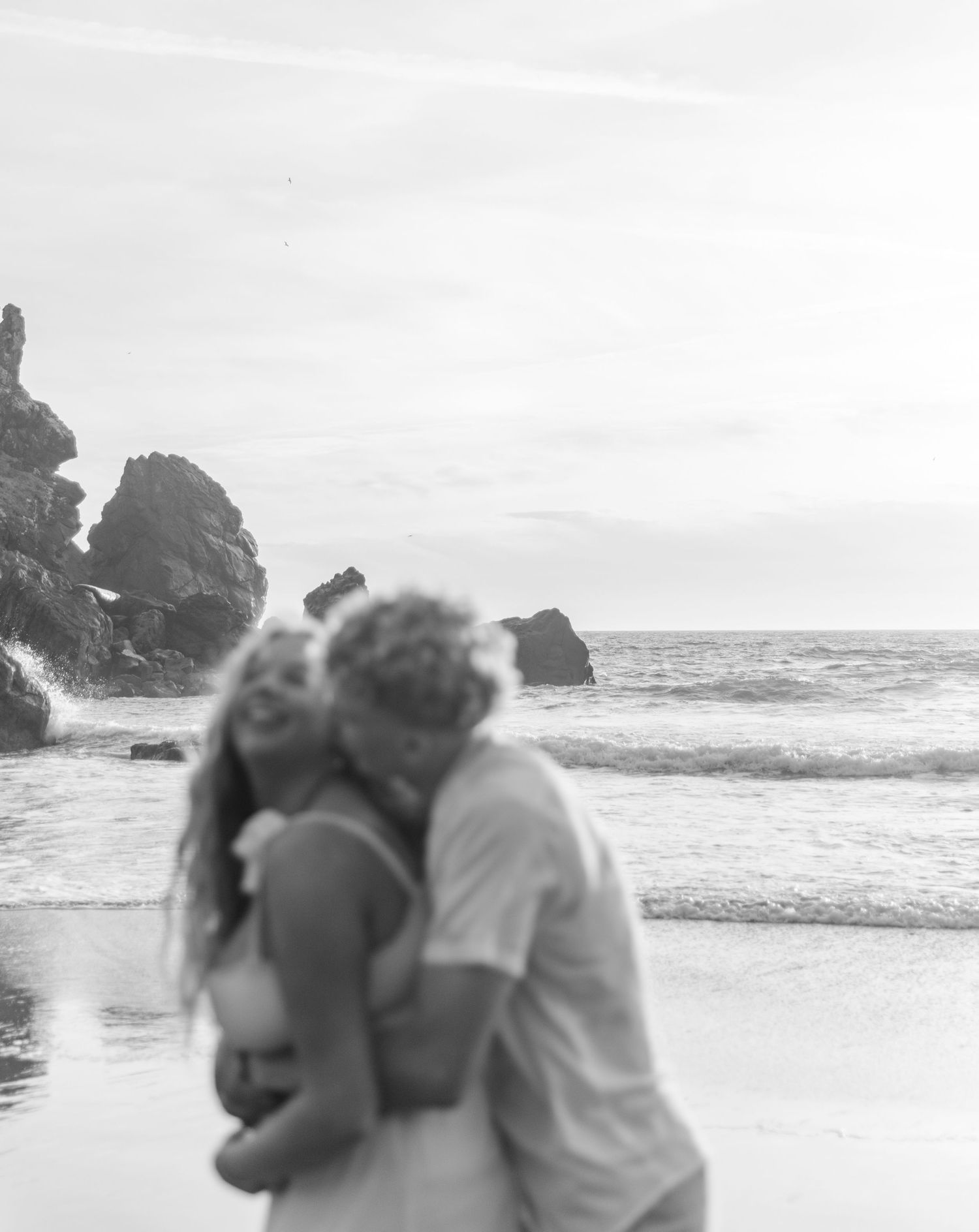 A couple embraces on a rocky beach with crashing waves and dramatic coastal cliffs in the background.