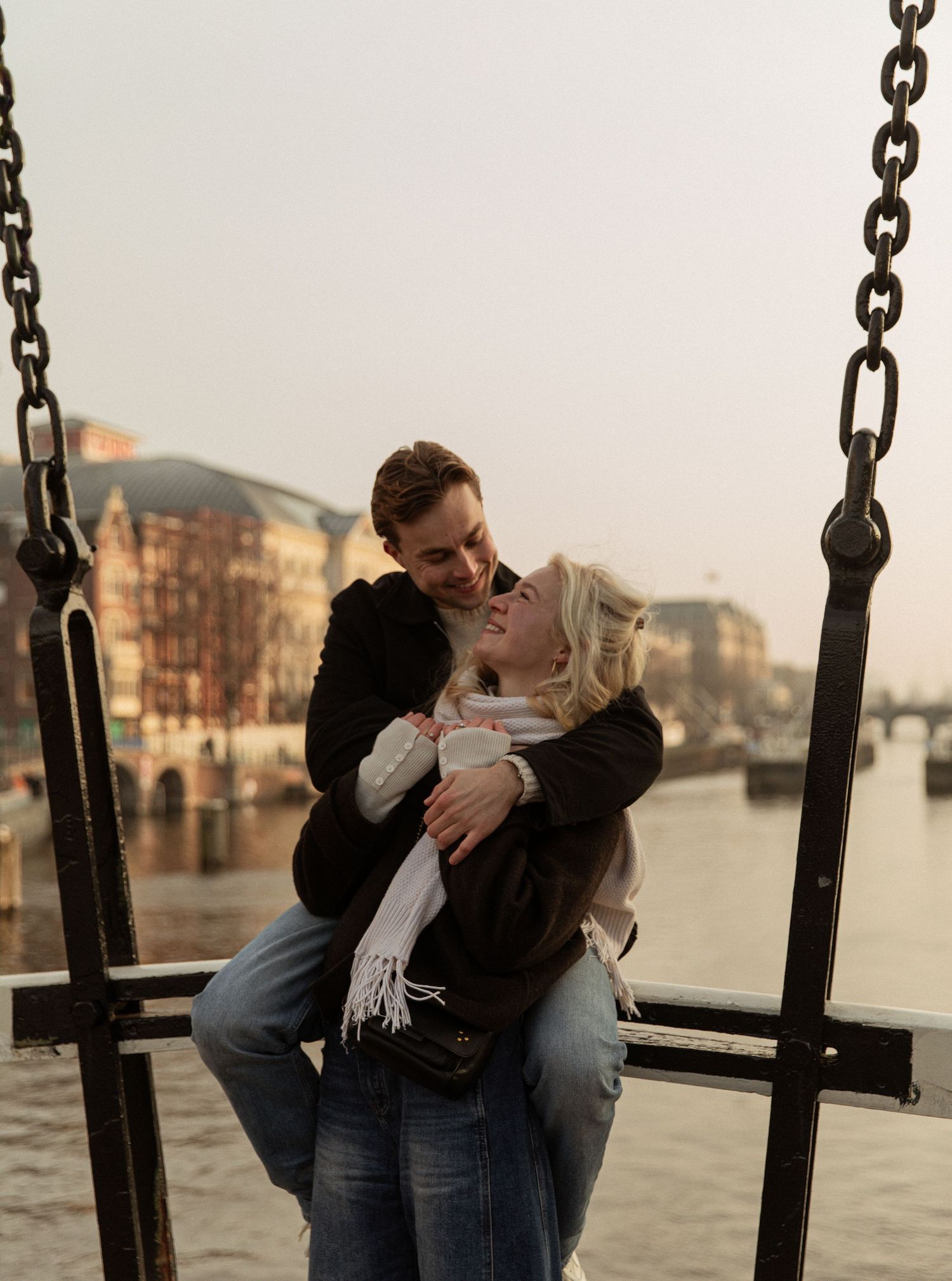 A couple shares an intimate moment while sitting on a swing by a waterfront at sunset.