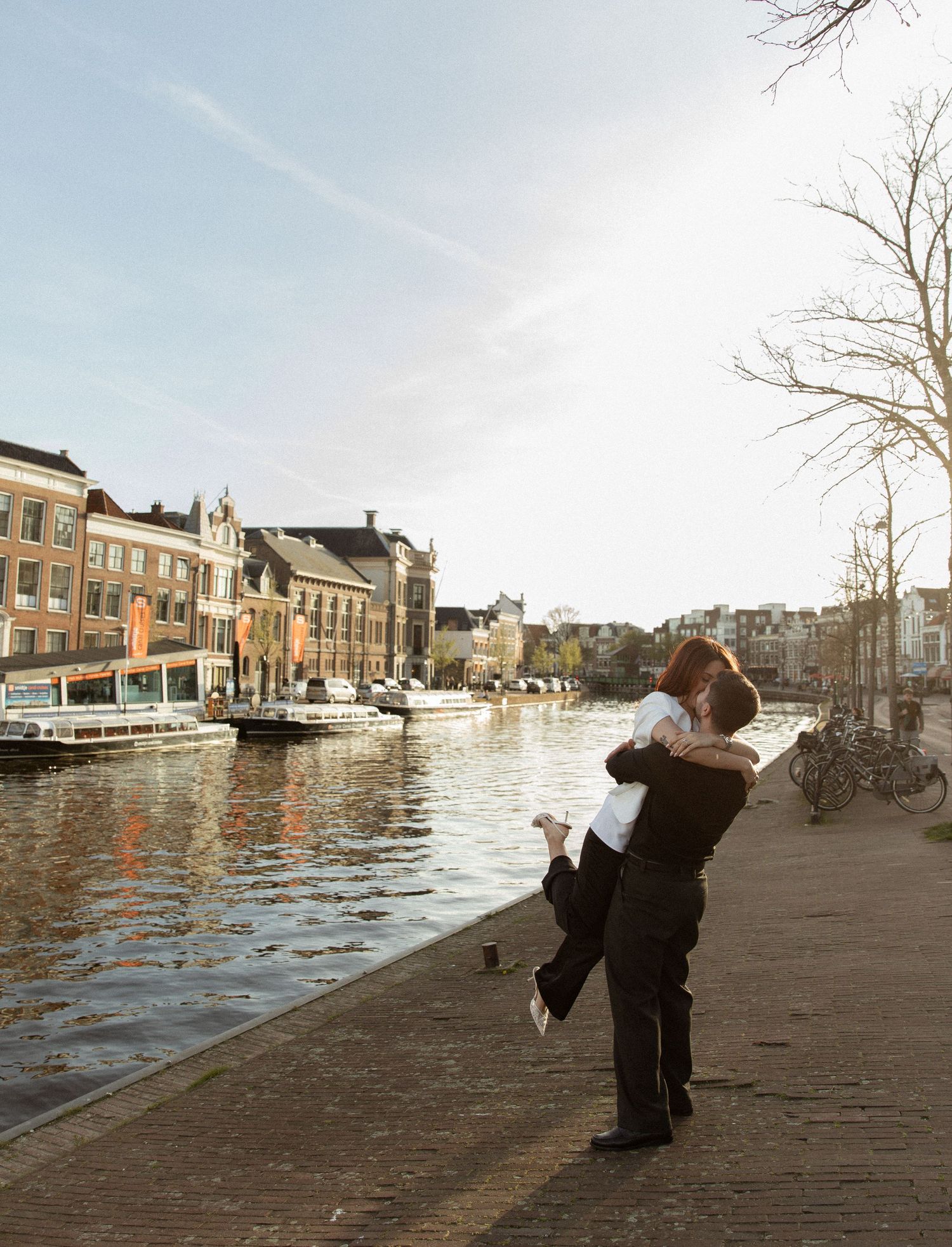 Couple embracing by Amsterdam canal on sunny day, with historic buildings and boats reflected in calm water.