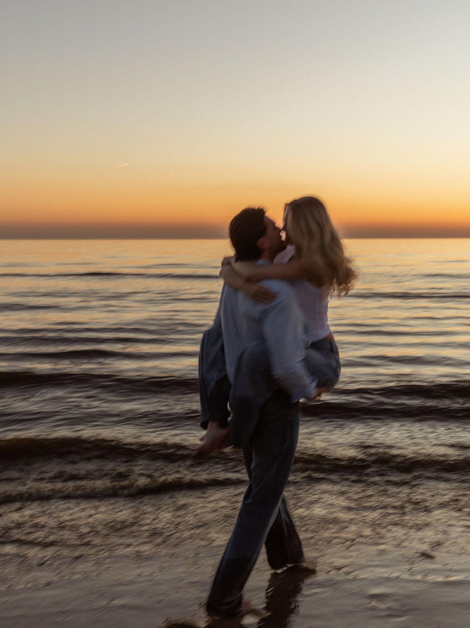 Couple kissing at sunset on a beach, man carrying woman in his arms with golden horizon over calm ocean.