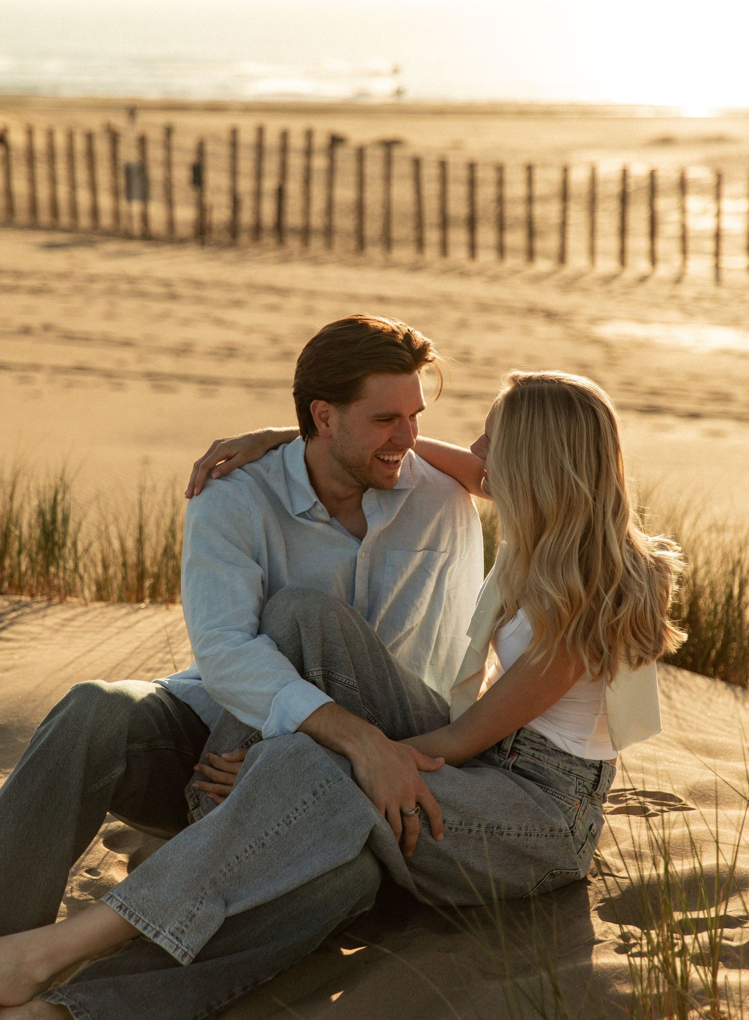Couple sitting together on sandy beach dunes at golden hour, smiling at each other near a wooden fence.
