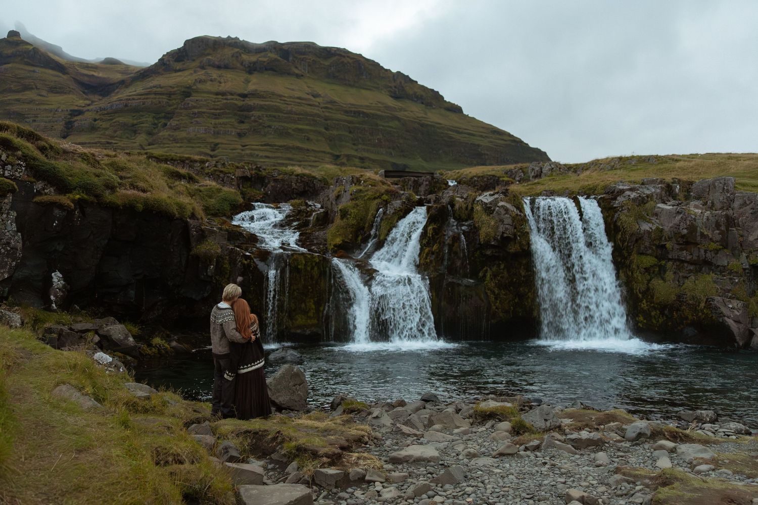 Person standing before cascading waterfalls with lush green hills in Iceland's dramatic landscape.