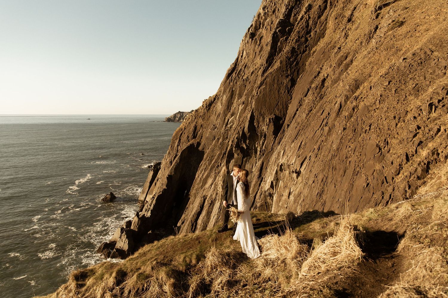 A person in white stands on a dramatic coastal cliff edge overlooking the Pacific Ocean on a misty day.