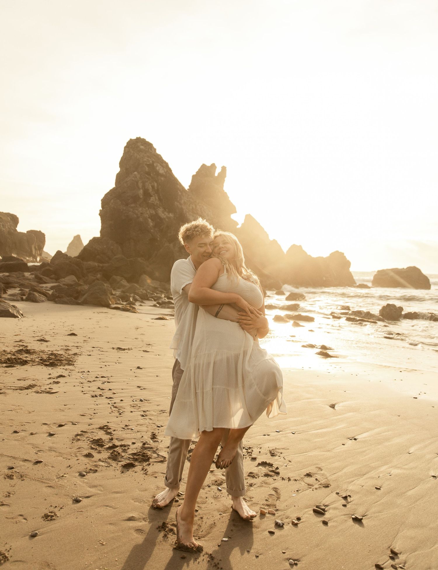 Romantic couple embraces on a sunset beach with rocky cliffs and golden light creating a dreamy silhouette.