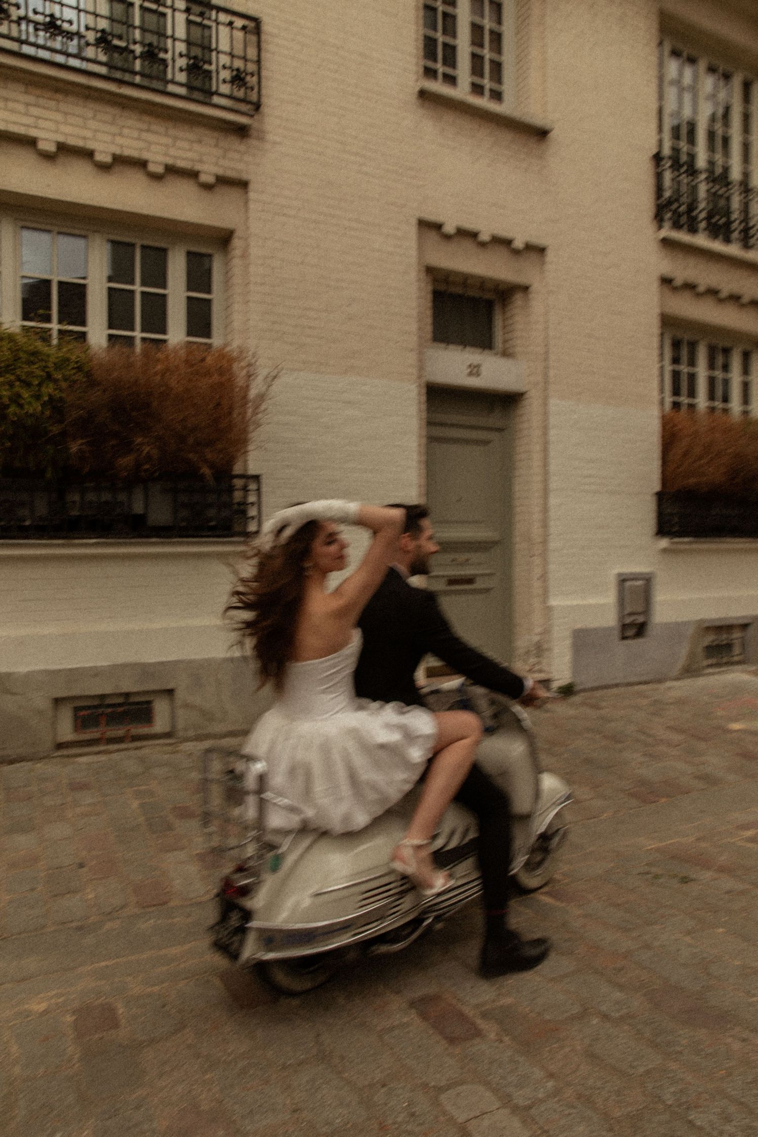 A bride and groom ride a vintage scooter through cobblestone Parisian streets, her white dress and hair flowing in motion blur.