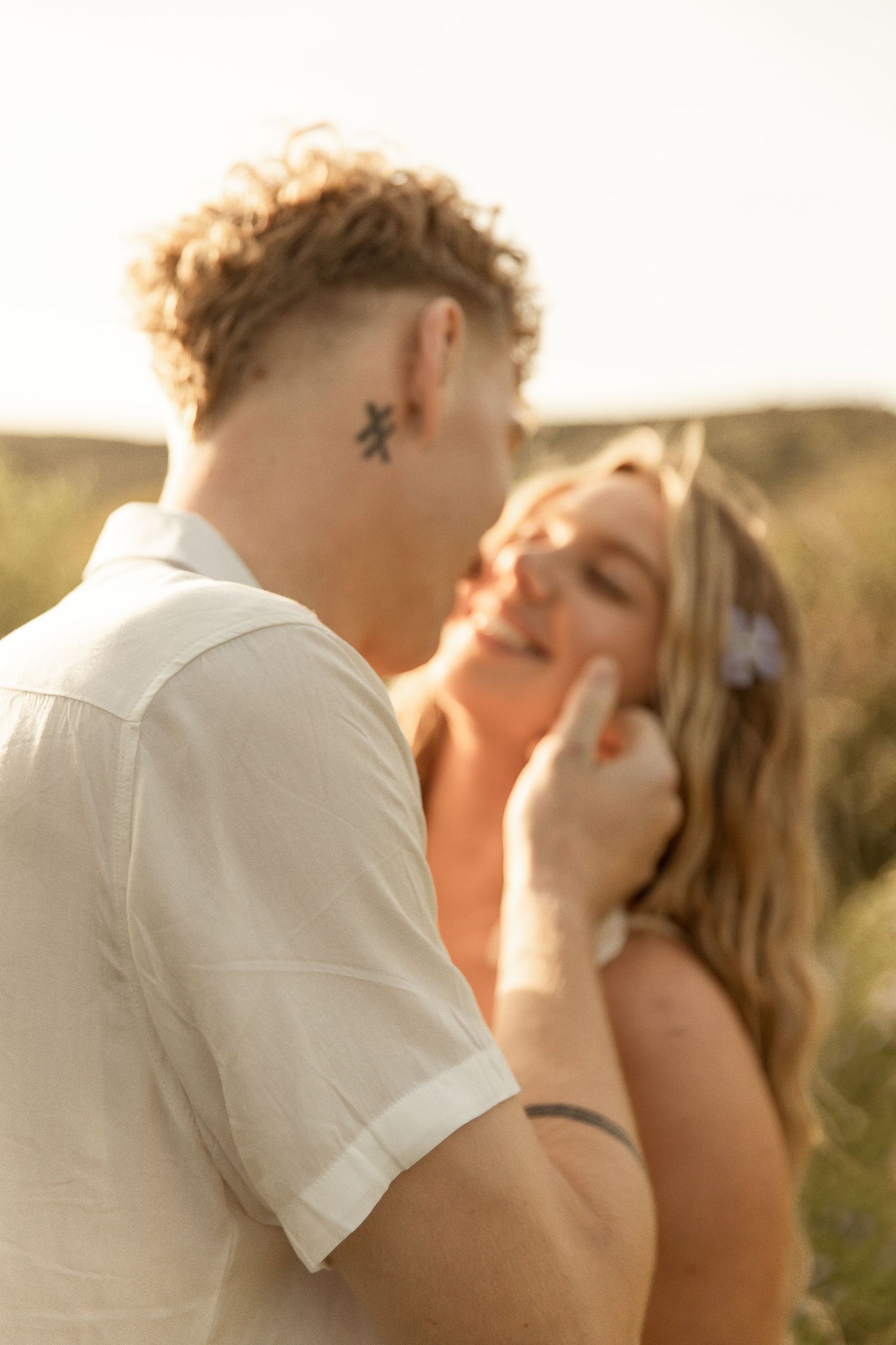 A romantic couple embraces and kisses during golden hour in a natural outdoor setting.