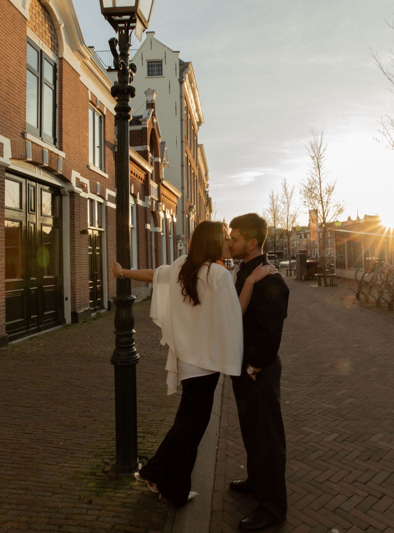 Couple sharing a romantic kiss by a lamppost on a cobblestone street at golden hour in a European city.