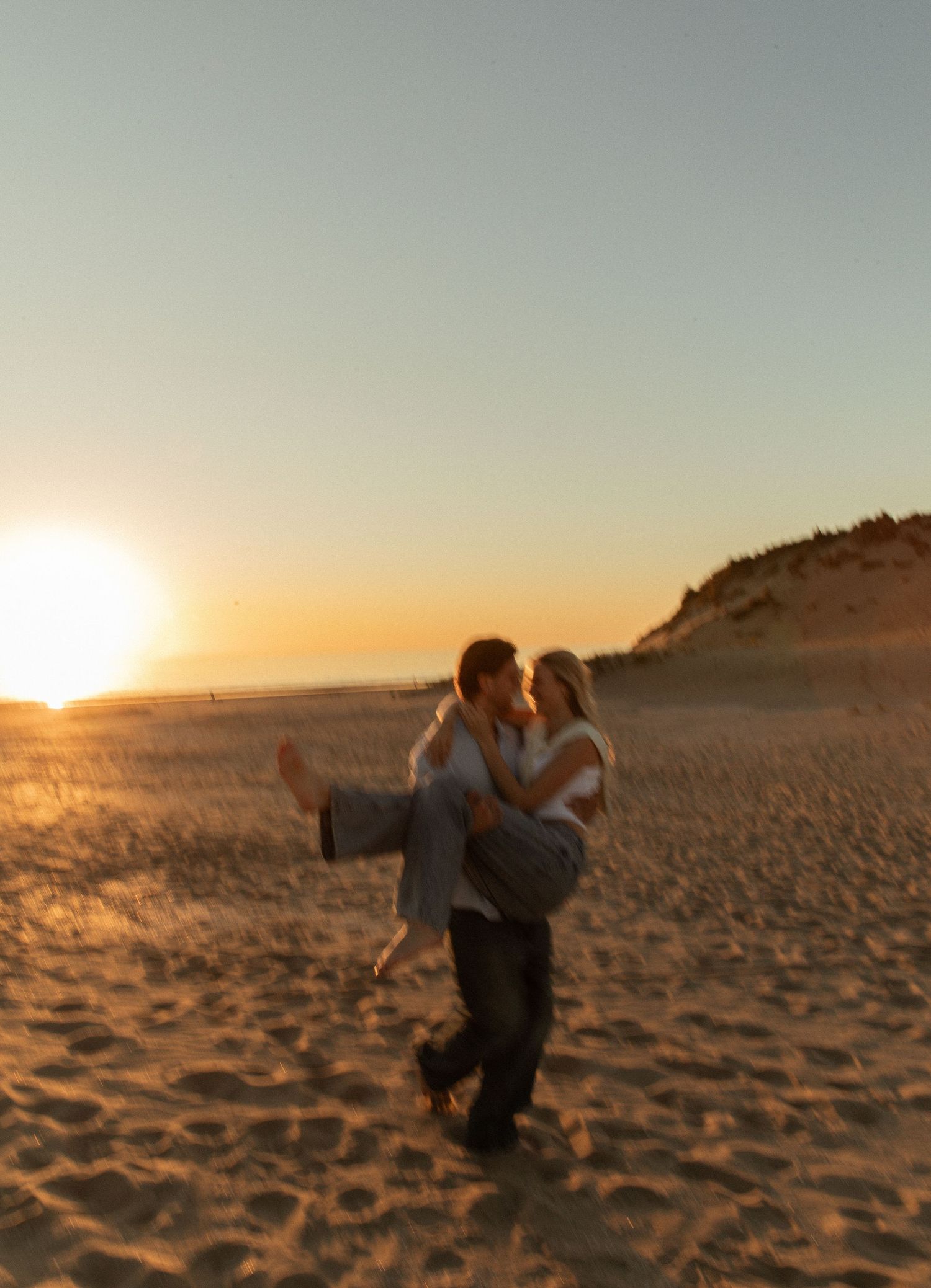 Couple sharing a romantic kiss at sunset on a sandy beach, man lifting woman in his arms.