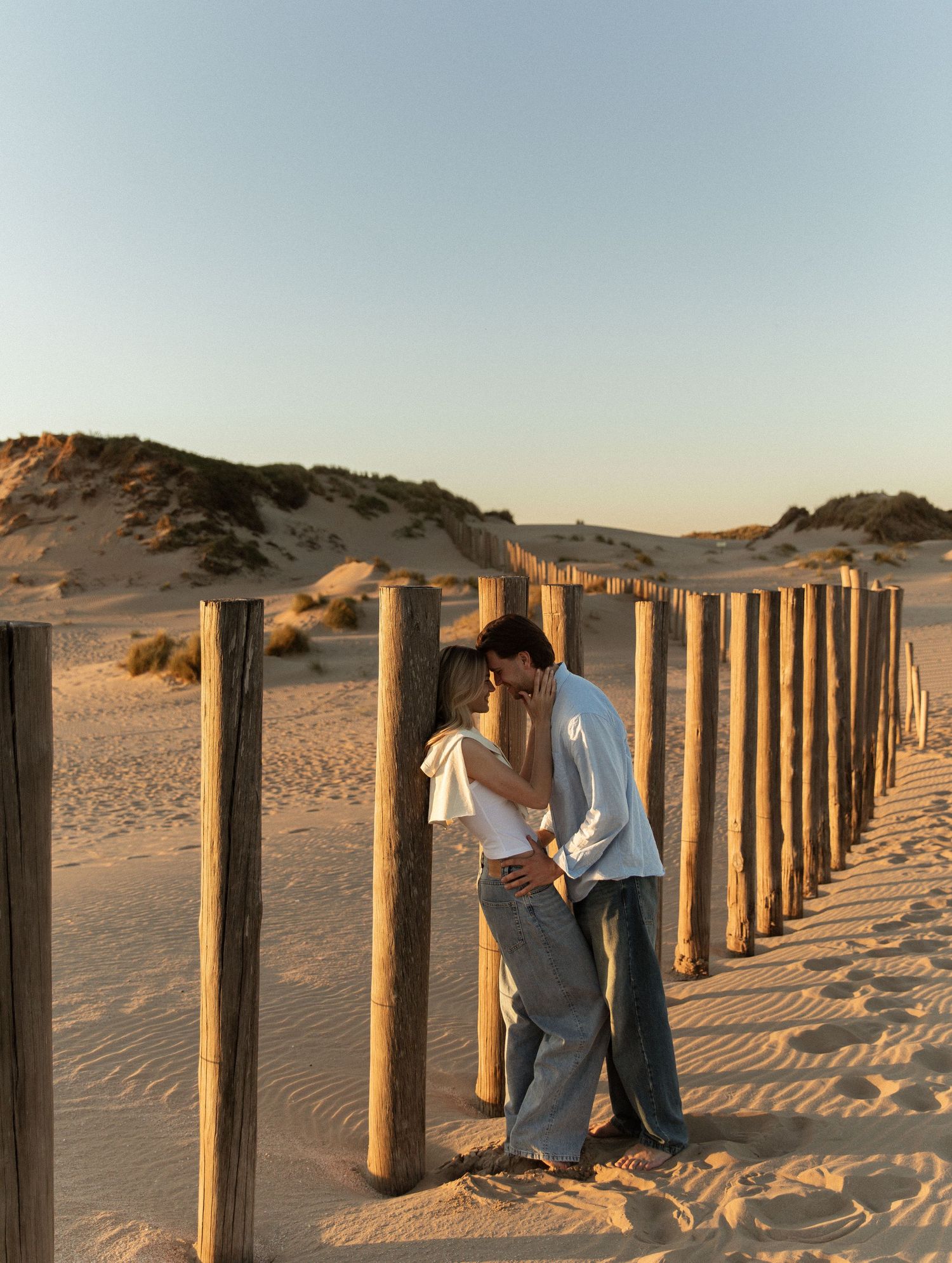 Couple kissing between wooden posts on a sandy beach at golden hour with dunes in the background.