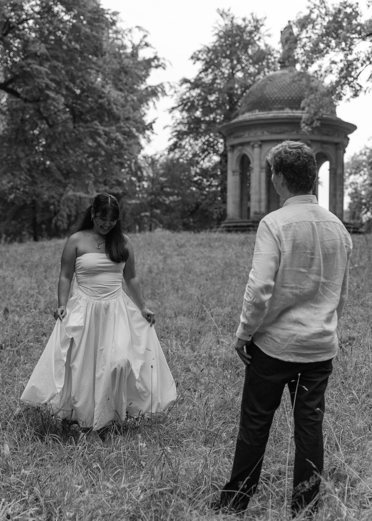 Black and white photo of a couple in wedding attire standing in a grassy field with a historic gazebo in the background.